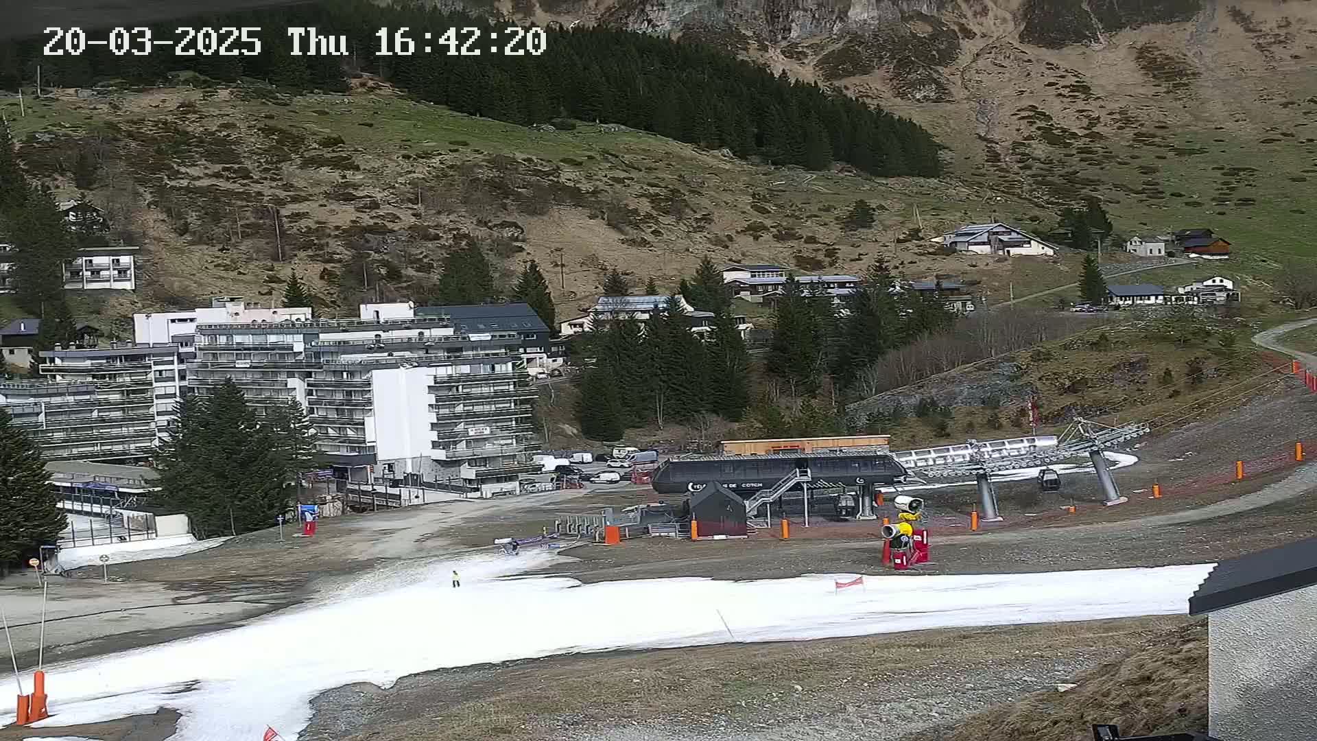A ski lift and several buildings are situated on a mountainside with patchy snow cover, under a partly cloudy sky.