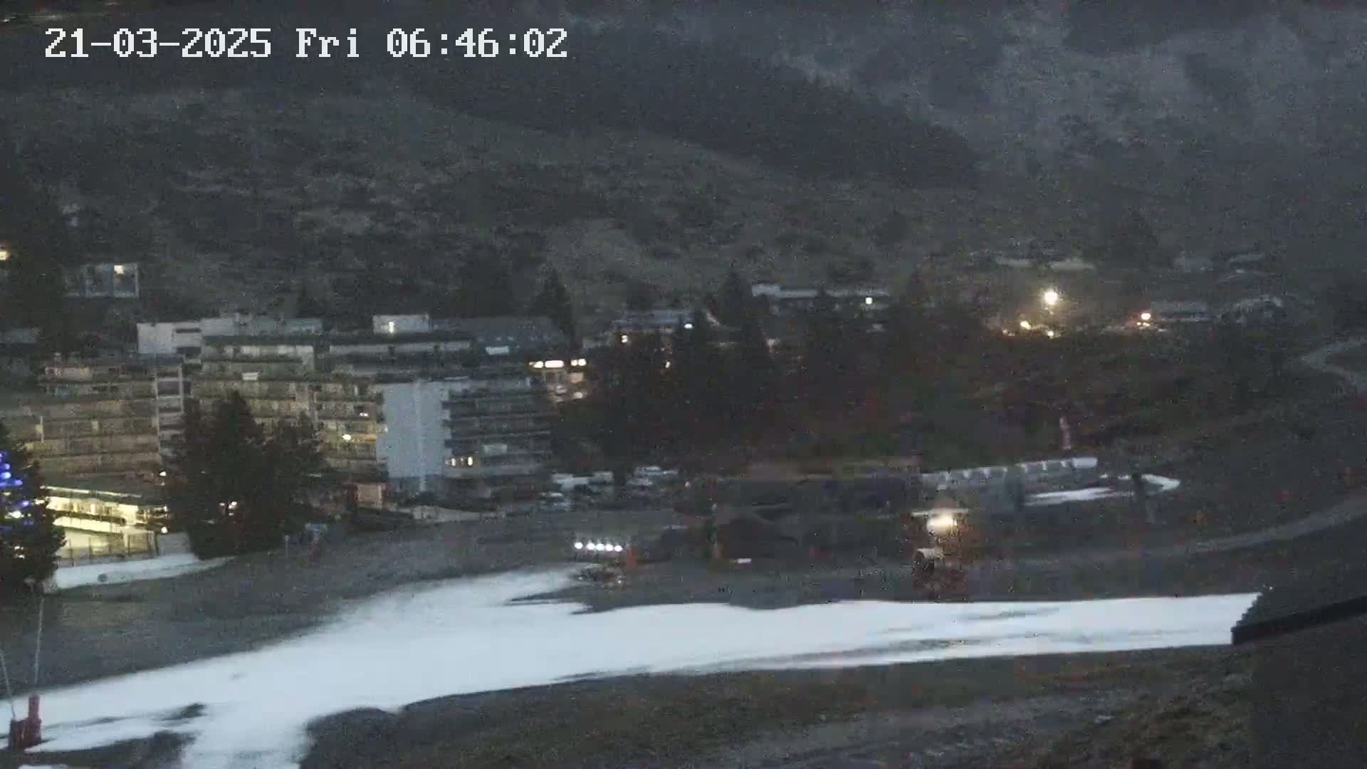 A snow-covered area in front of a building complex at the base of a dark mountain under a lightly snowing, dusky sky.
