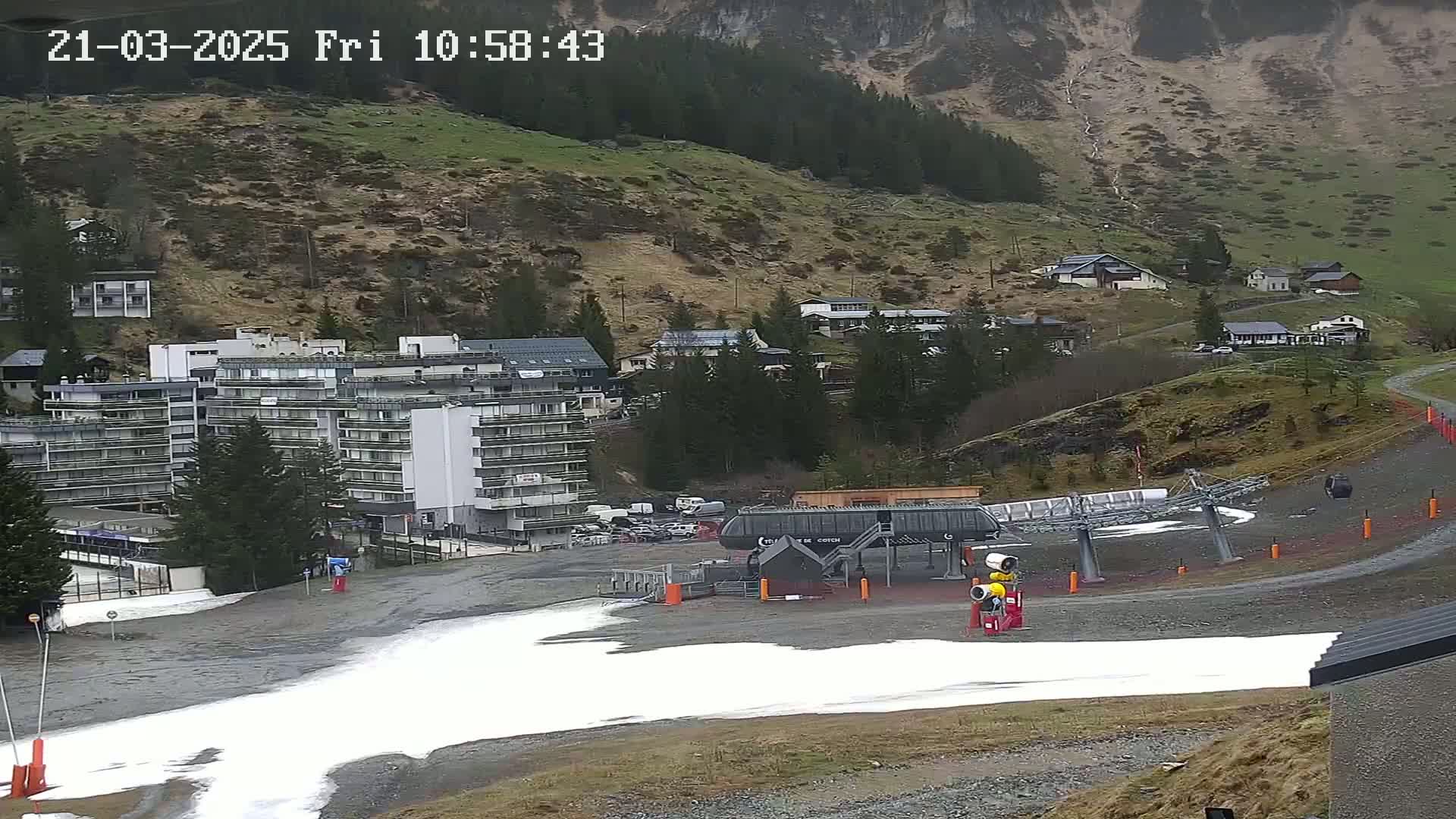 A ski lift base station with patchy snow sits in a valley below a hillside of brown grass and dark trees under an overcast sky.