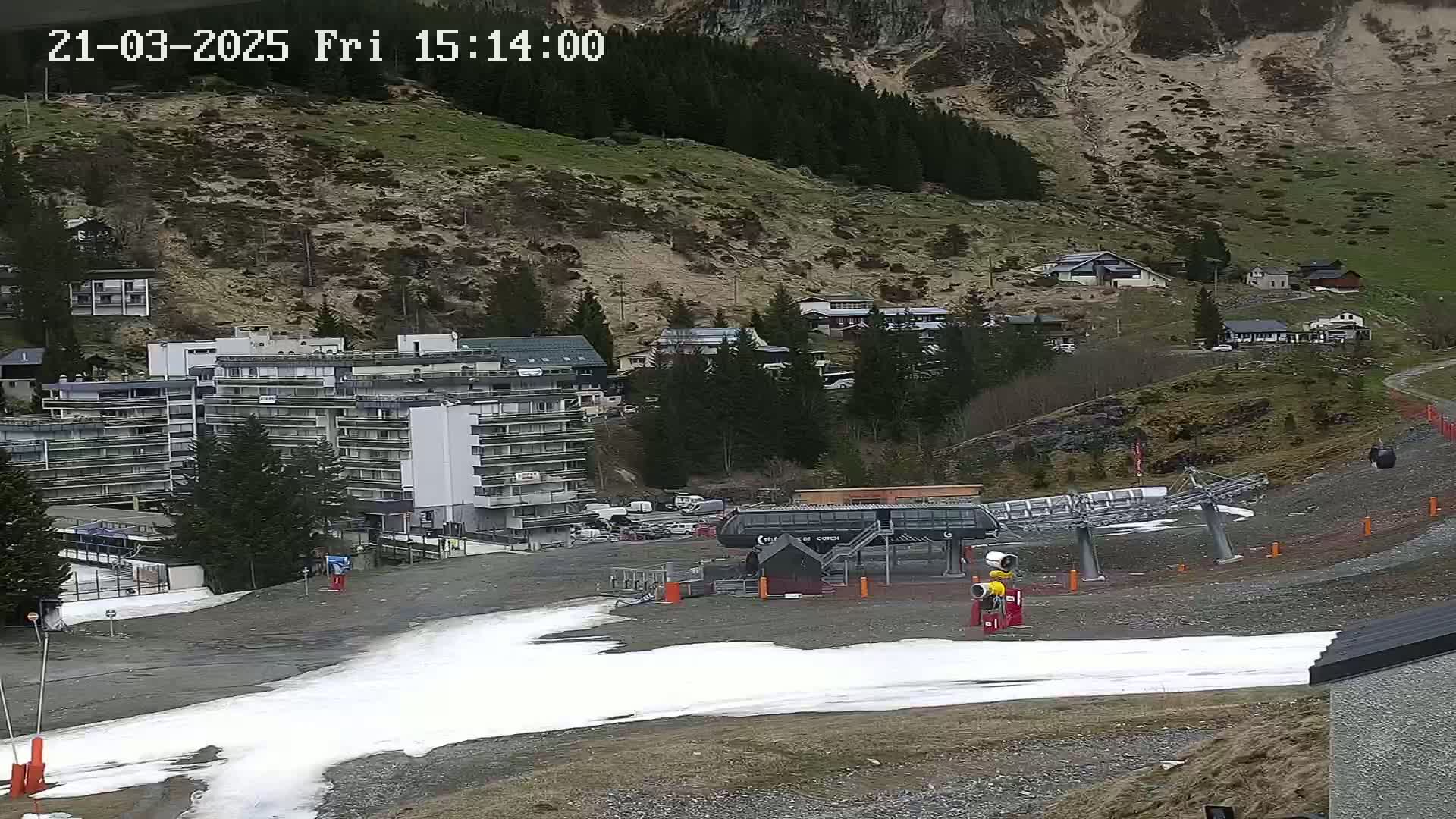 A chairlift and a multi-story building are situated at the base of a mountain, with patches of snow on the ground in an overcast setting.