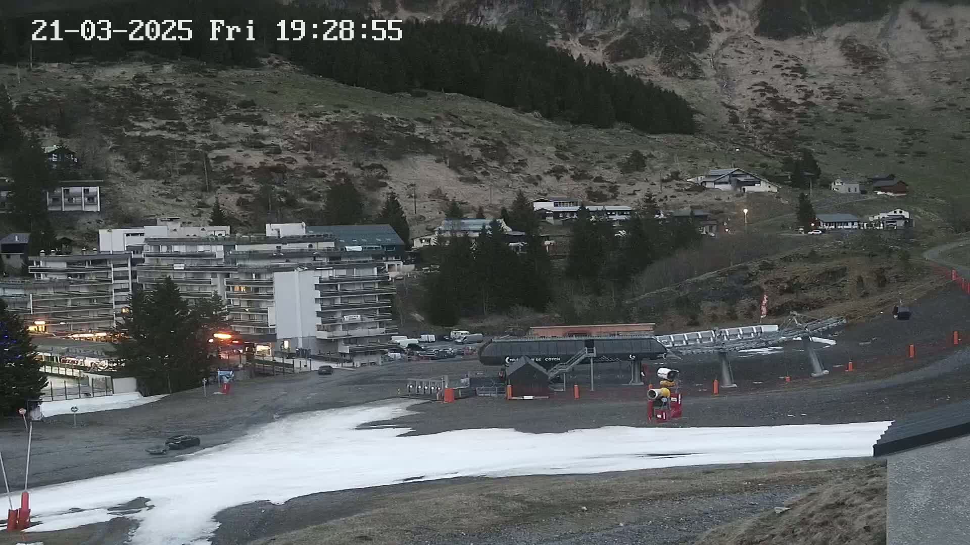 A ski lift base station sits in a mountainside village at dusk, with patches of melting snow visible on the ground.
