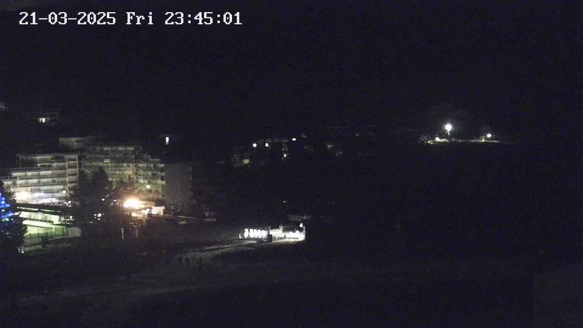 A nighttime view shows a multi-story building and other structures, dimly lit, with a dark, possibly sandy or paved area in the foreground.