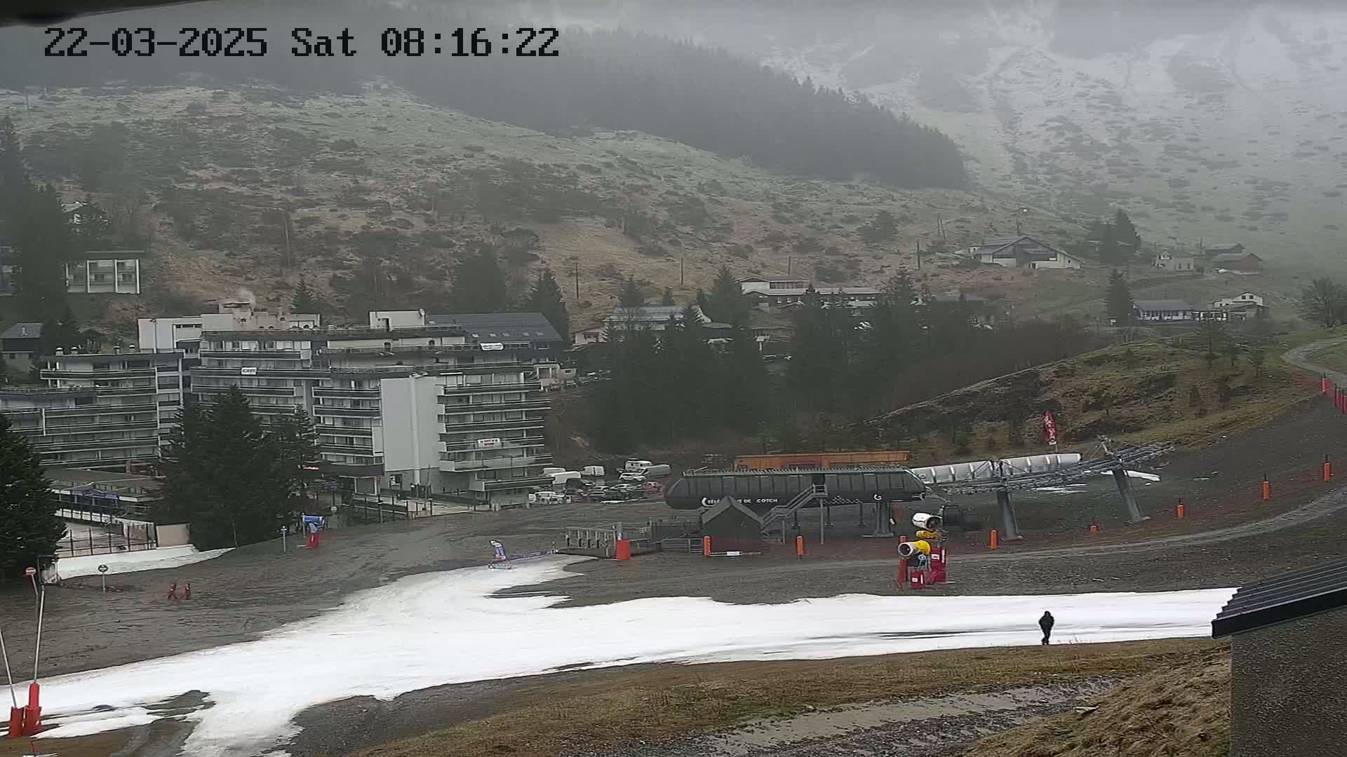 A ski lift base area with a multi-story building in the background on a misty, overcast day with patches of snow on the ground.
