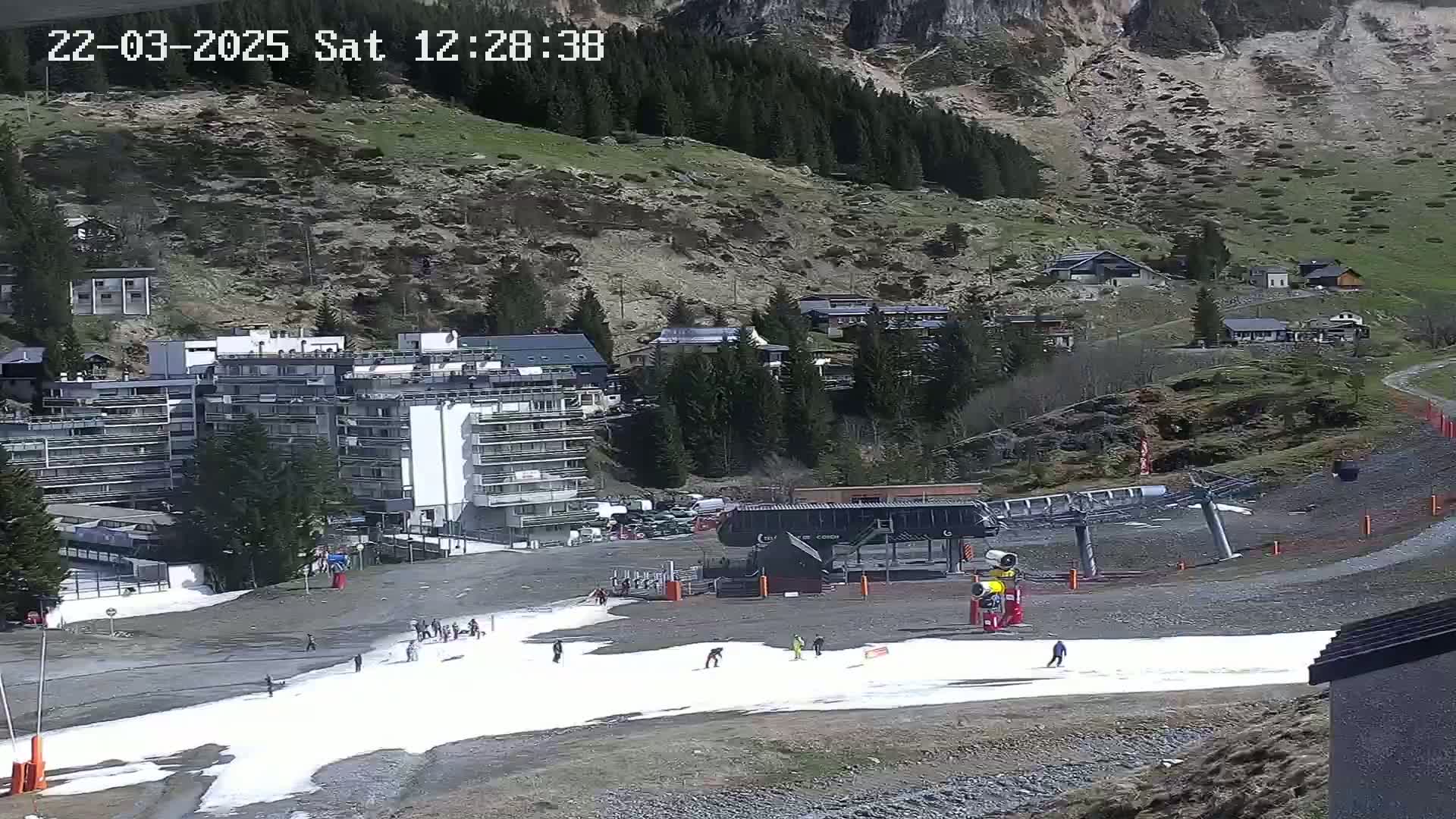 A ski resort scene shows a mostly-empty ski slope with patches of melting snow, a ski lift, several buildings, and a mountainside with sparse vegetation in the background, under sunny conditions.