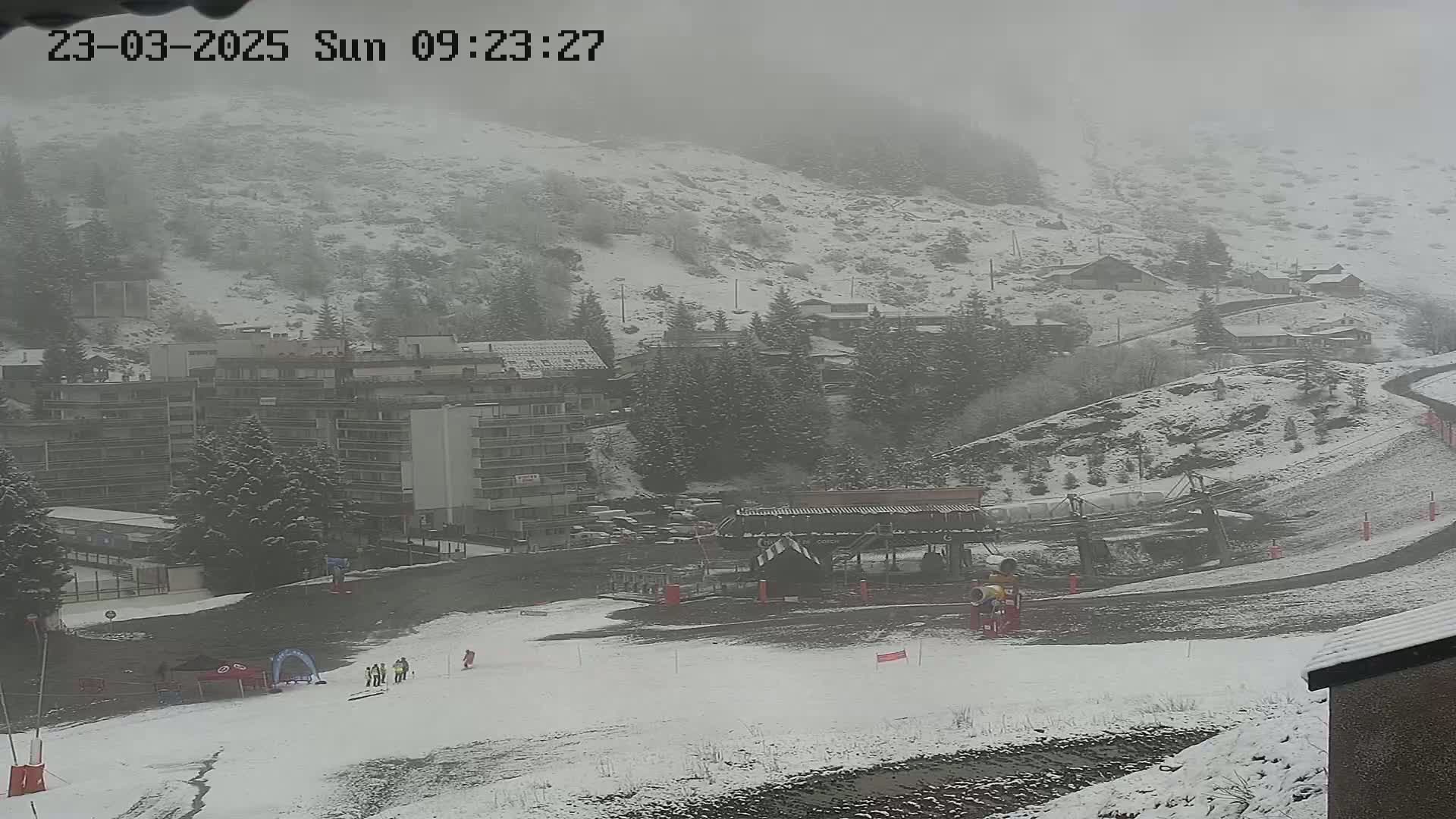 A snow-covered ski resort is seen in foggy conditions, with a ski lift and buildings visible in the distance.