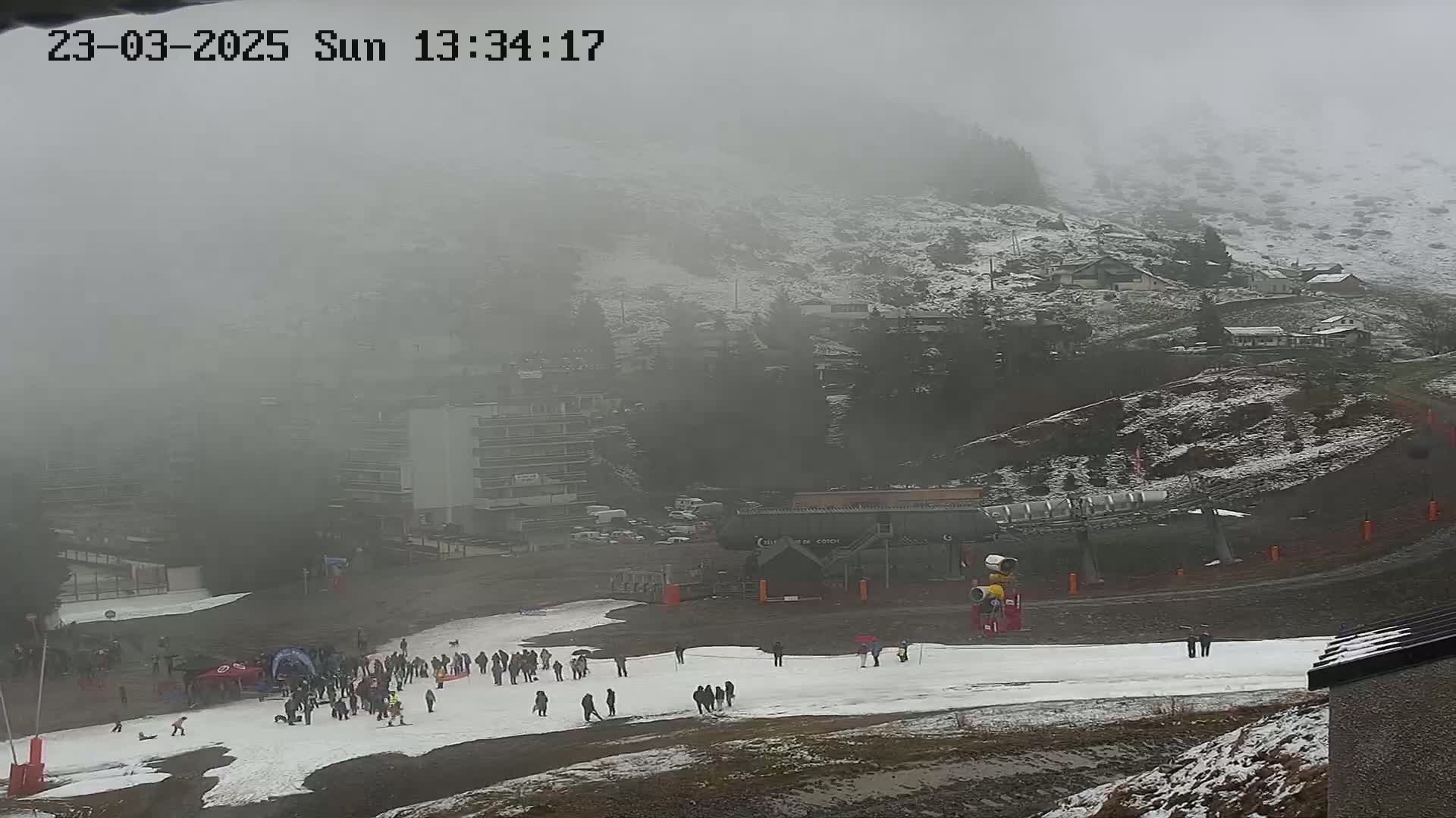 A foggy, snowy mountain scene shows a ski resort with a chairlift, several buildings, and a scattering of people on a snowy slope.
