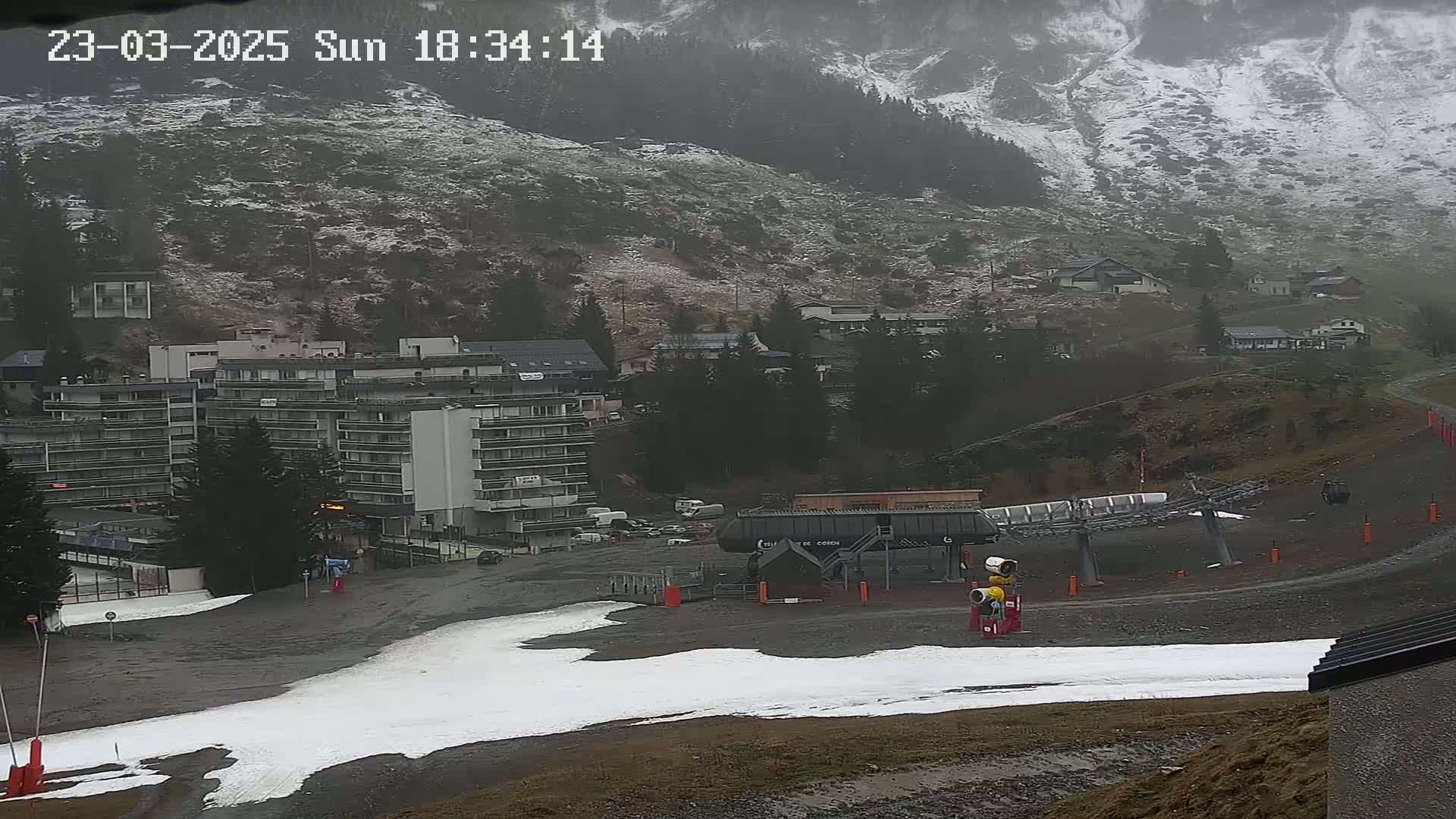 A snow-dusted mountainside overlooks a ski resort with a chairlift and several buildings, under overcast, snowy weather.