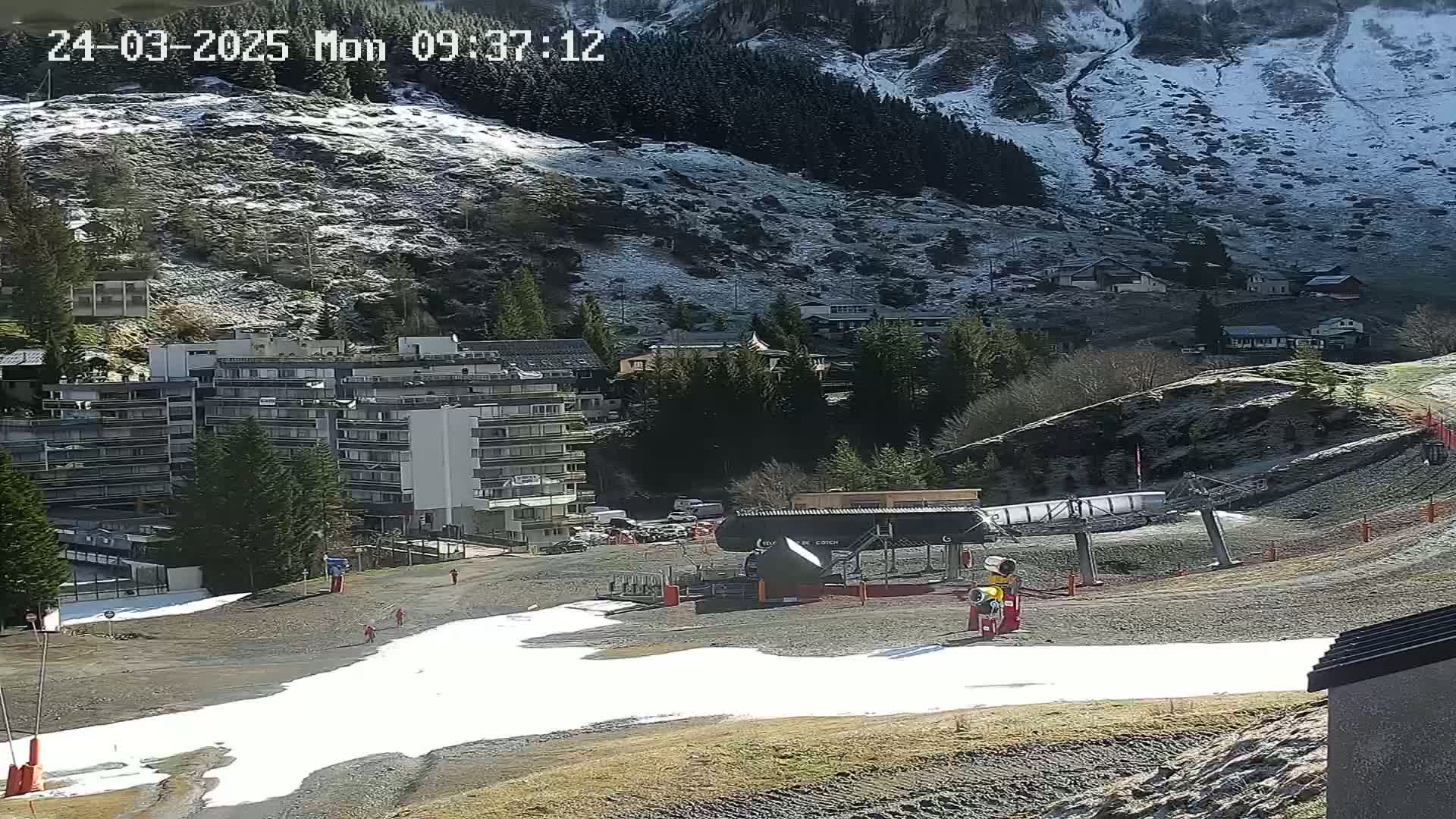 A ski lift and snowmaking equipment sit in a mountain valley on a partly sunny day with patches of snow and ice on the ground and mountainside.