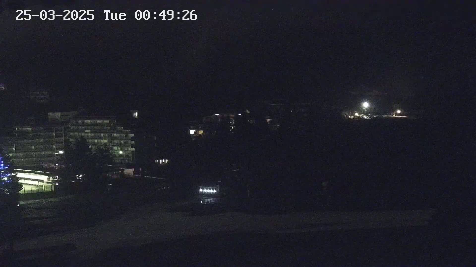 A dark nighttime scene shows a multi-story building and other structures illuminated by a few lights, with a possibly snowy ground in the foreground.