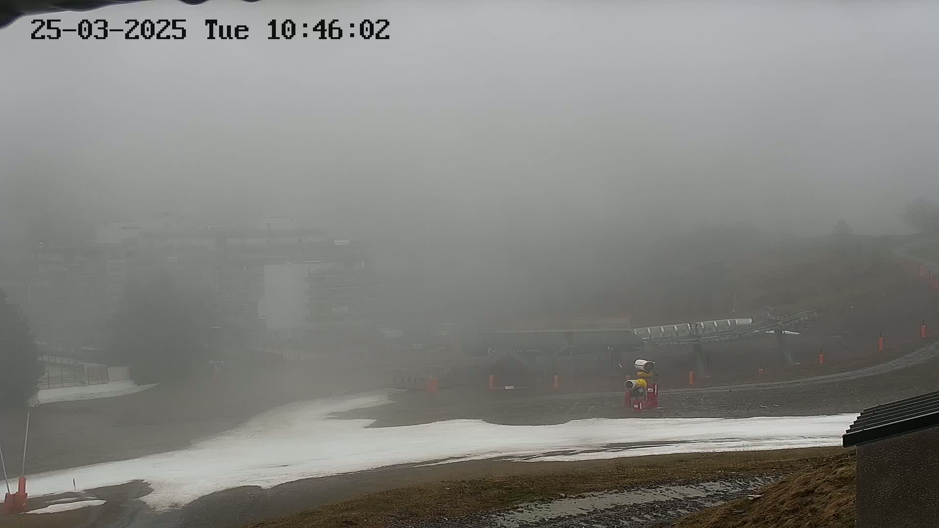 A foggy view of a ski slope with a chairlift and buildings barely visible in the distance.