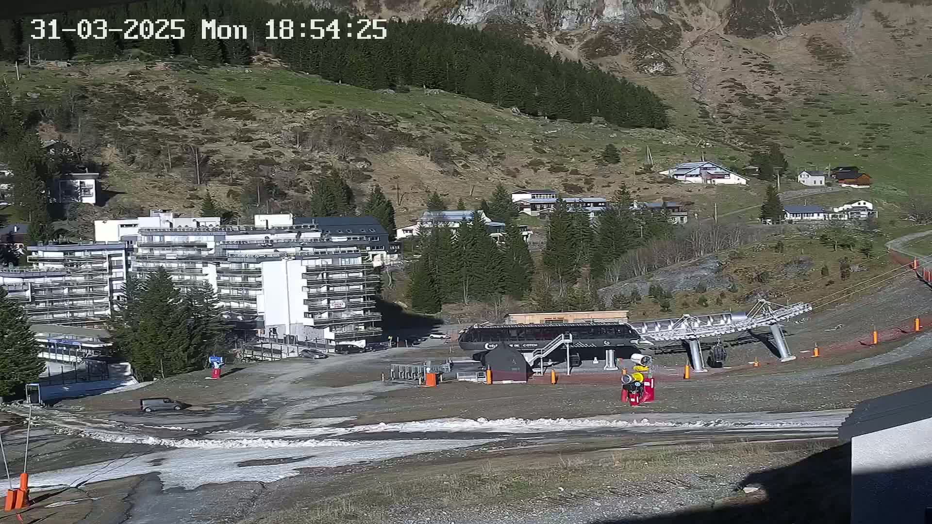 A ski lift base station and a large apartment building are situated in a mountain village on a sunny day with patches of melting snow.