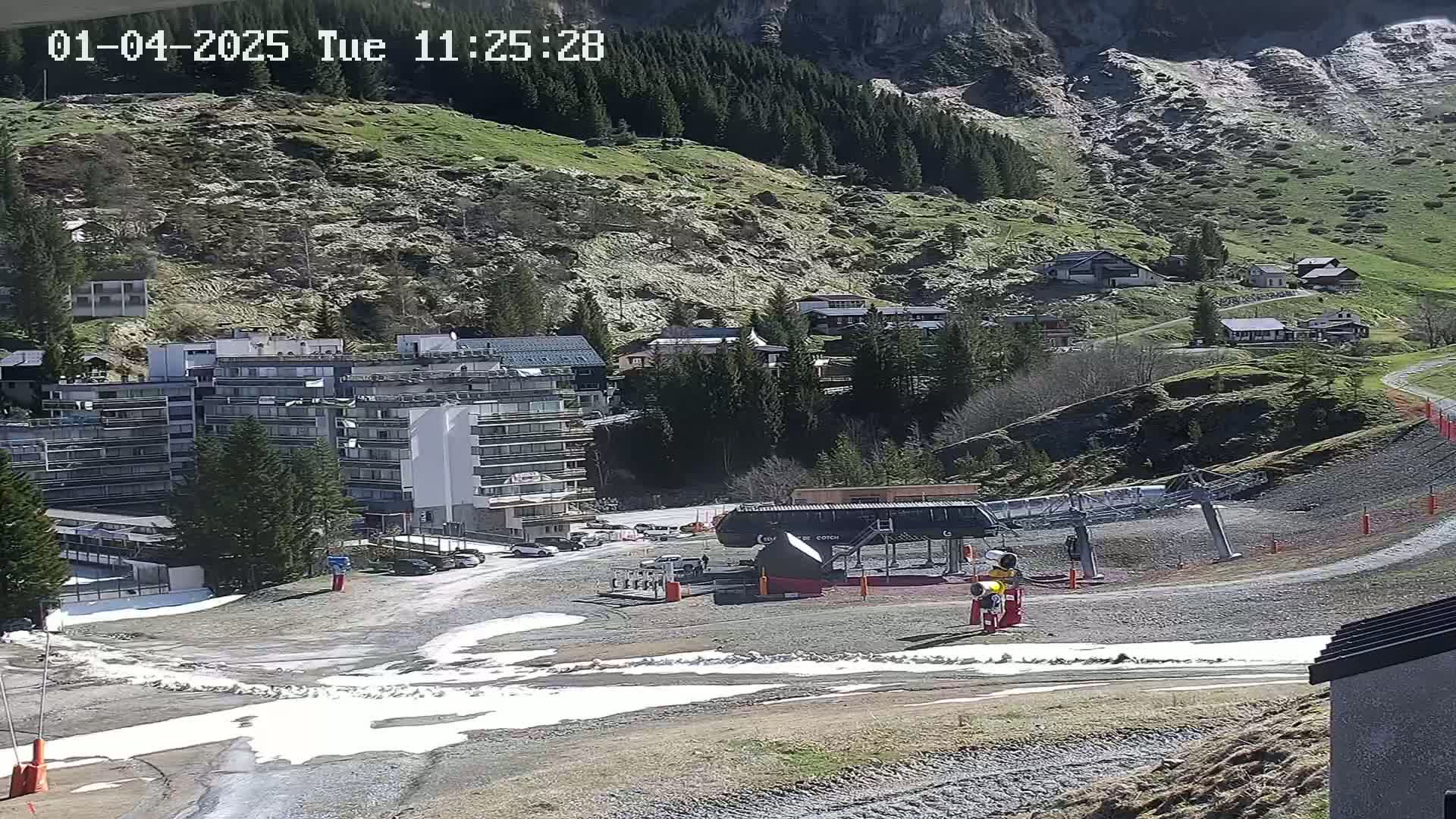 A mountainside resort town with patches of snow and a chairlift visible on a sunny day.