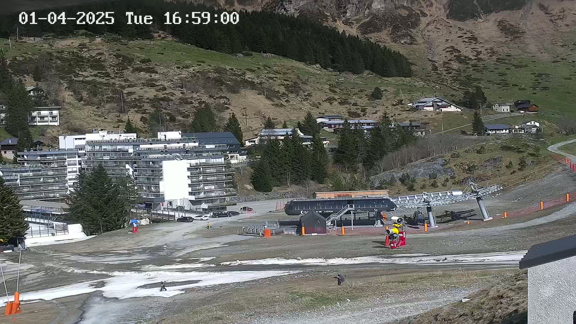 A ski lift base station sits in a mountain valley next to a large apartment building, with patches of melting snow and a few people visible on a mostly clear day.