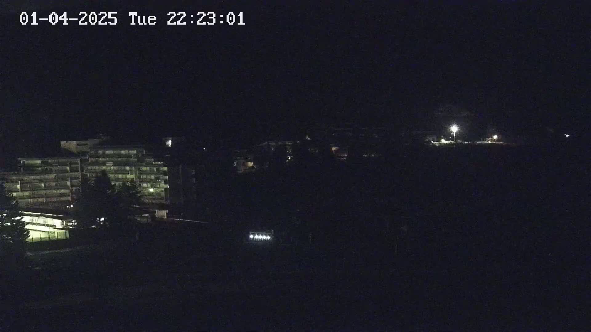 A nighttime long shot shows a multi-story building and other structures faintly illuminated under a dark, clear sky.