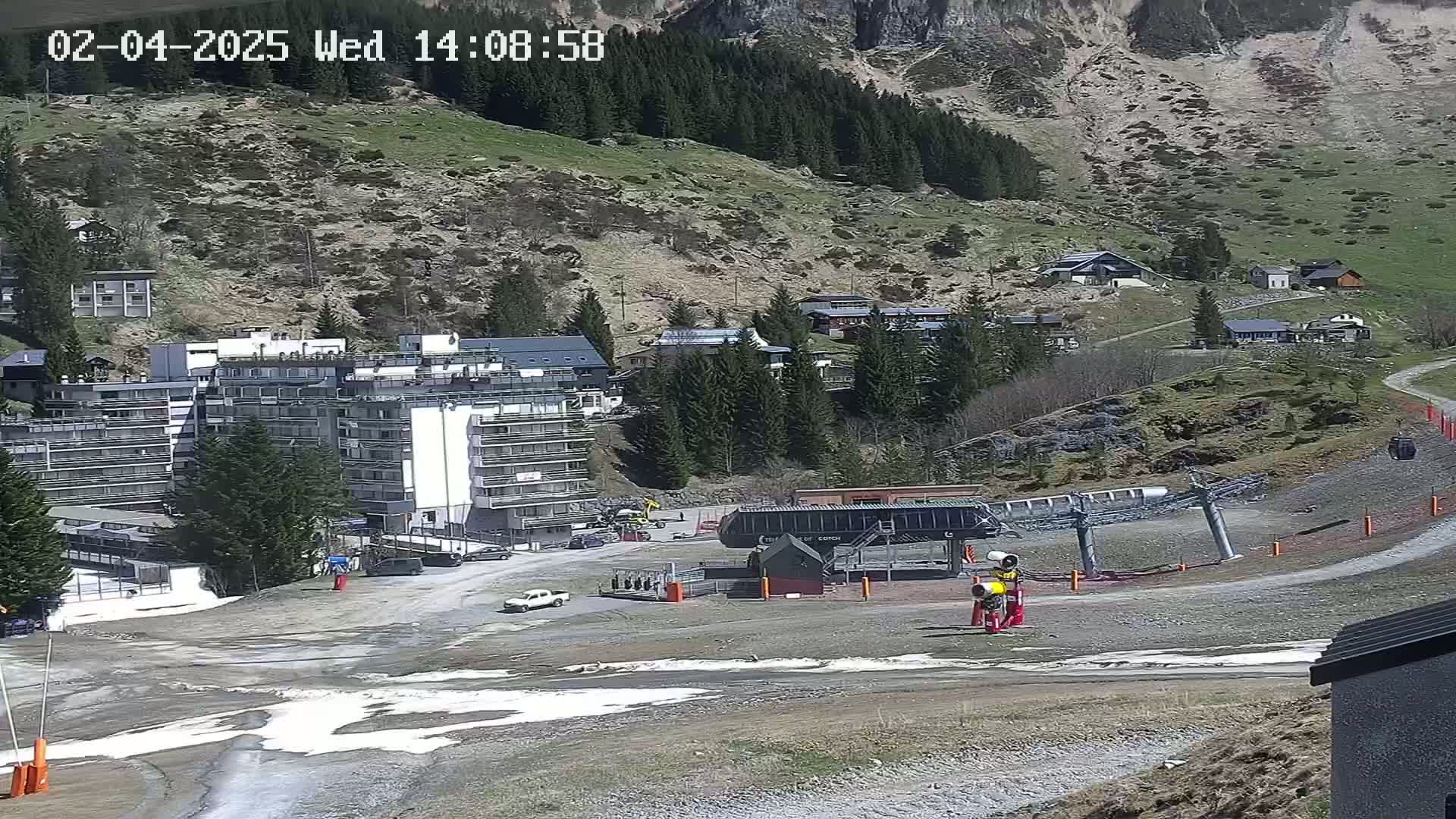 A ski lift base station and several buildings are situated on a mountainside with patchy snow and mostly clear weather.