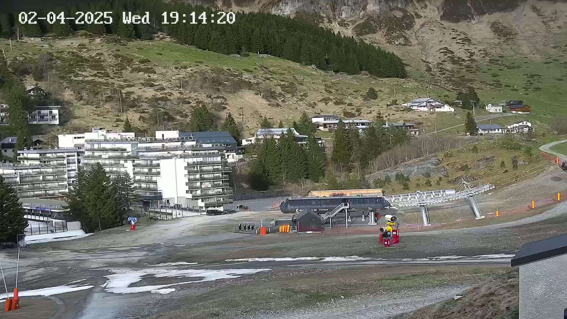A ski lift base station sits in a mountain village with patches of melting snow on the ground under a mostly clear sky.