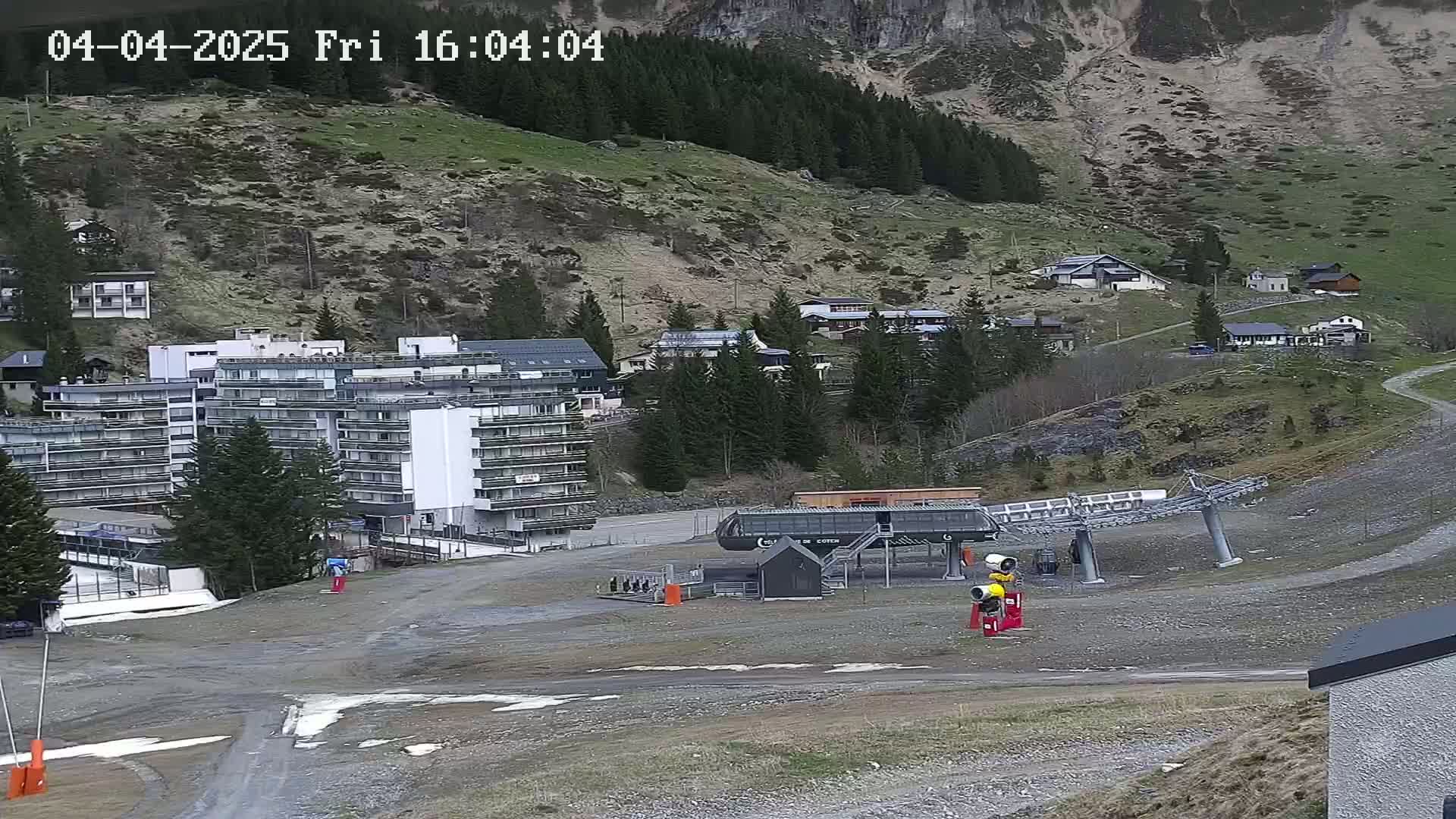 A ski lift base station sits in a gravel lot below a large apartment building, nestled in a mountain valley on a partly cloudy day.