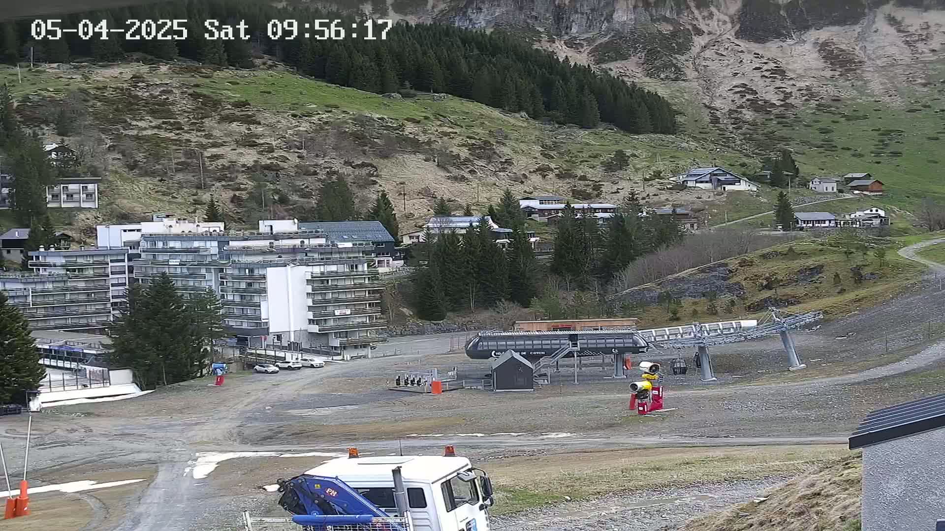 A ski lift base station is situated in a mountain valley, with a large building and several smaller houses visible, under a mostly clear sky.