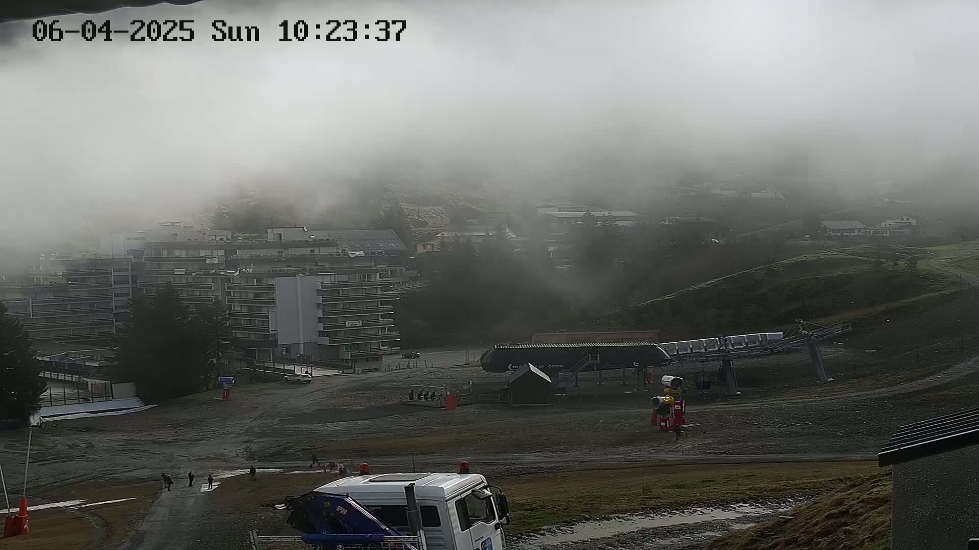 A foggy mountain scene shows a large apartment building, a ski lift, and a few people walking on a muddy area in front of a truck.