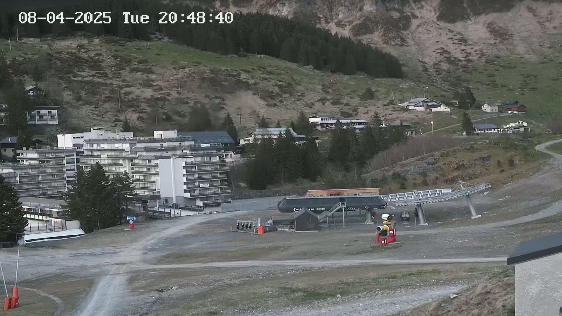 A ski lift base station sits in a gravel lot near a large apartment building and a hillside village, under twilight conditions.
