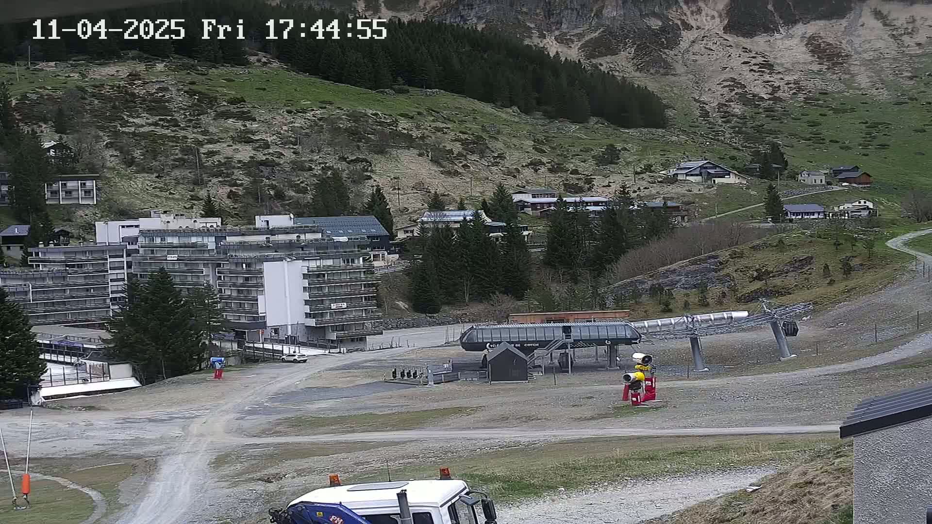 A ski lift base station and several buildings are situated on a gravel area at the base of a mountain with sparse trees and patchy grass under an overcast sky.