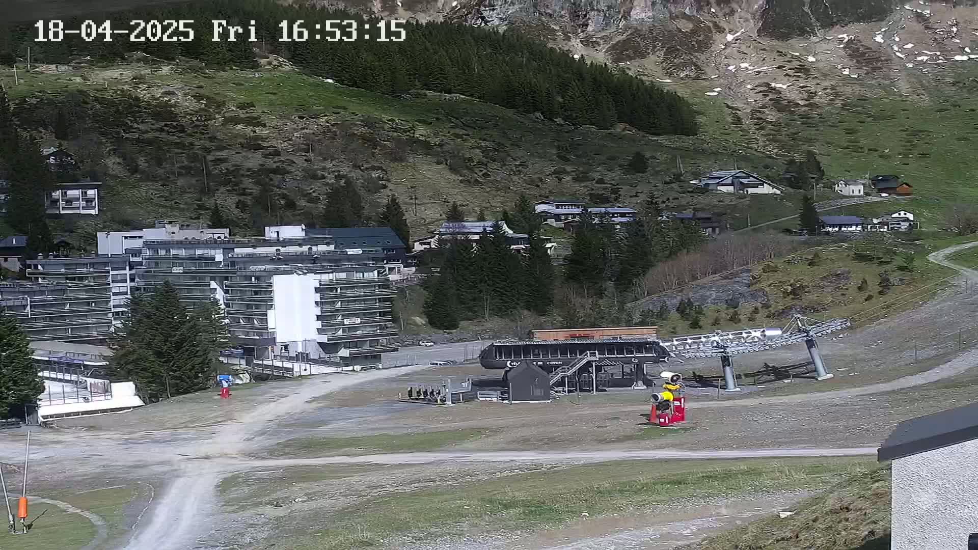 A sunny mountainside view shows a ski lift and several buildings, with a mostly barren, dirt ground in the foreground.
