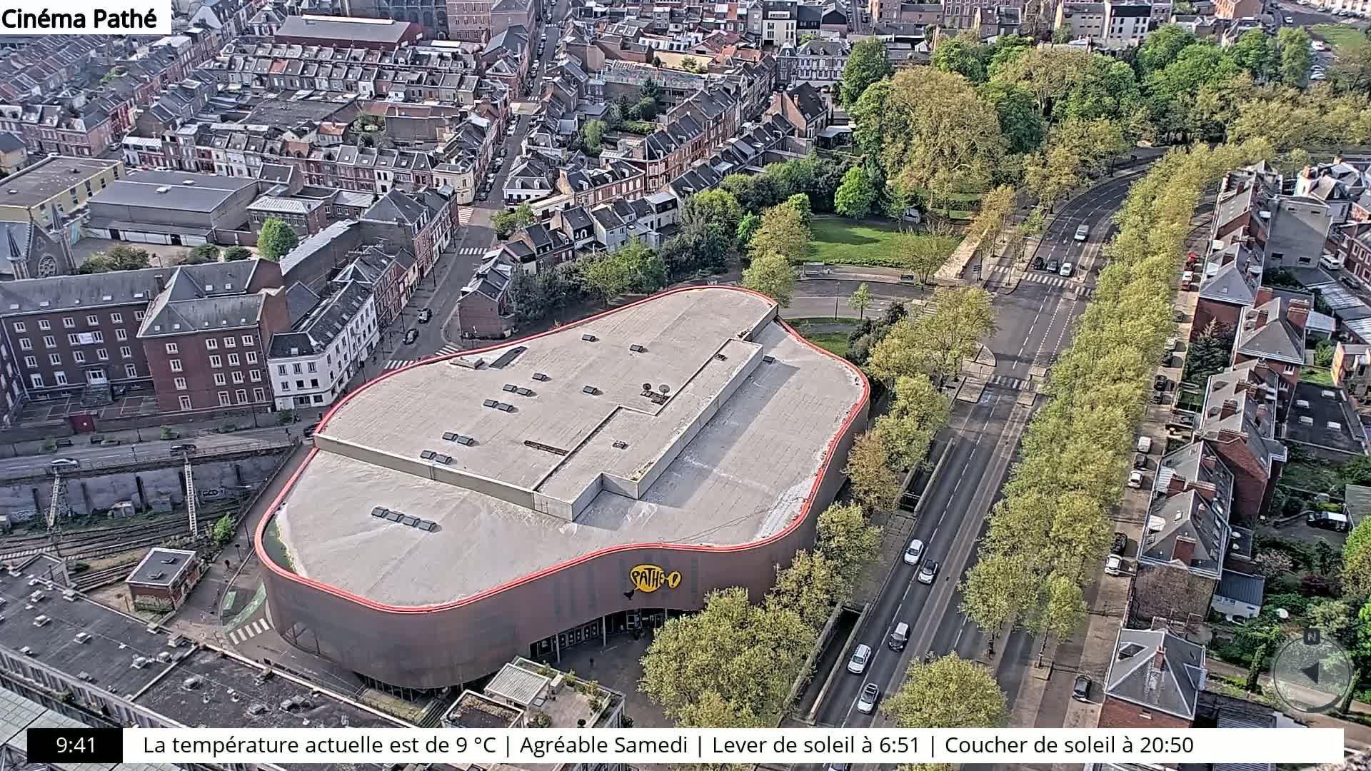 An aerial view shows a cinema building with a unique curved roof, surrounded by city streets and buildings, under a sunny sky.