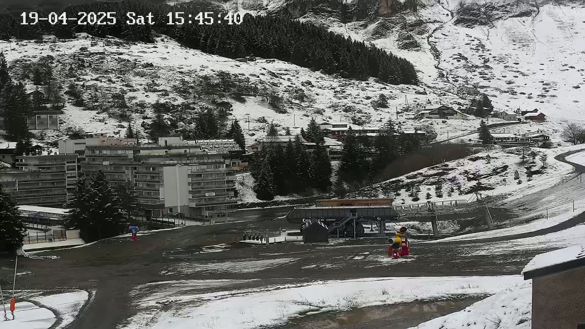A snow-dusted mountainside overlooks a village with buildings and a ski lift base, with patches of snow and muddy areas visible.