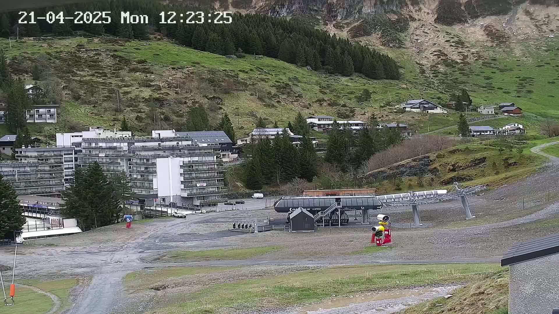 A ski lift base station and a large apartment building are situated in a mountainous area with patchy green grass and some trees, under overcast skies.