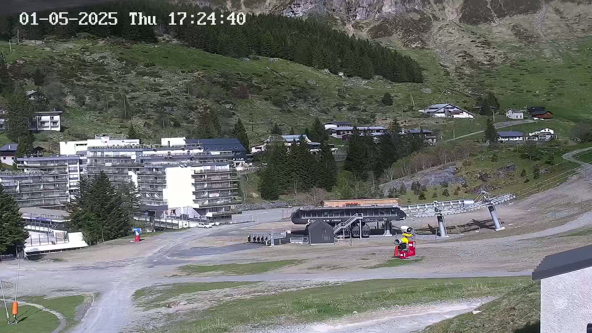 A mountainside view shows a chairlift base station, a large white apartment building, several smaller buildings, and green grassy hills under a sunny sky.
