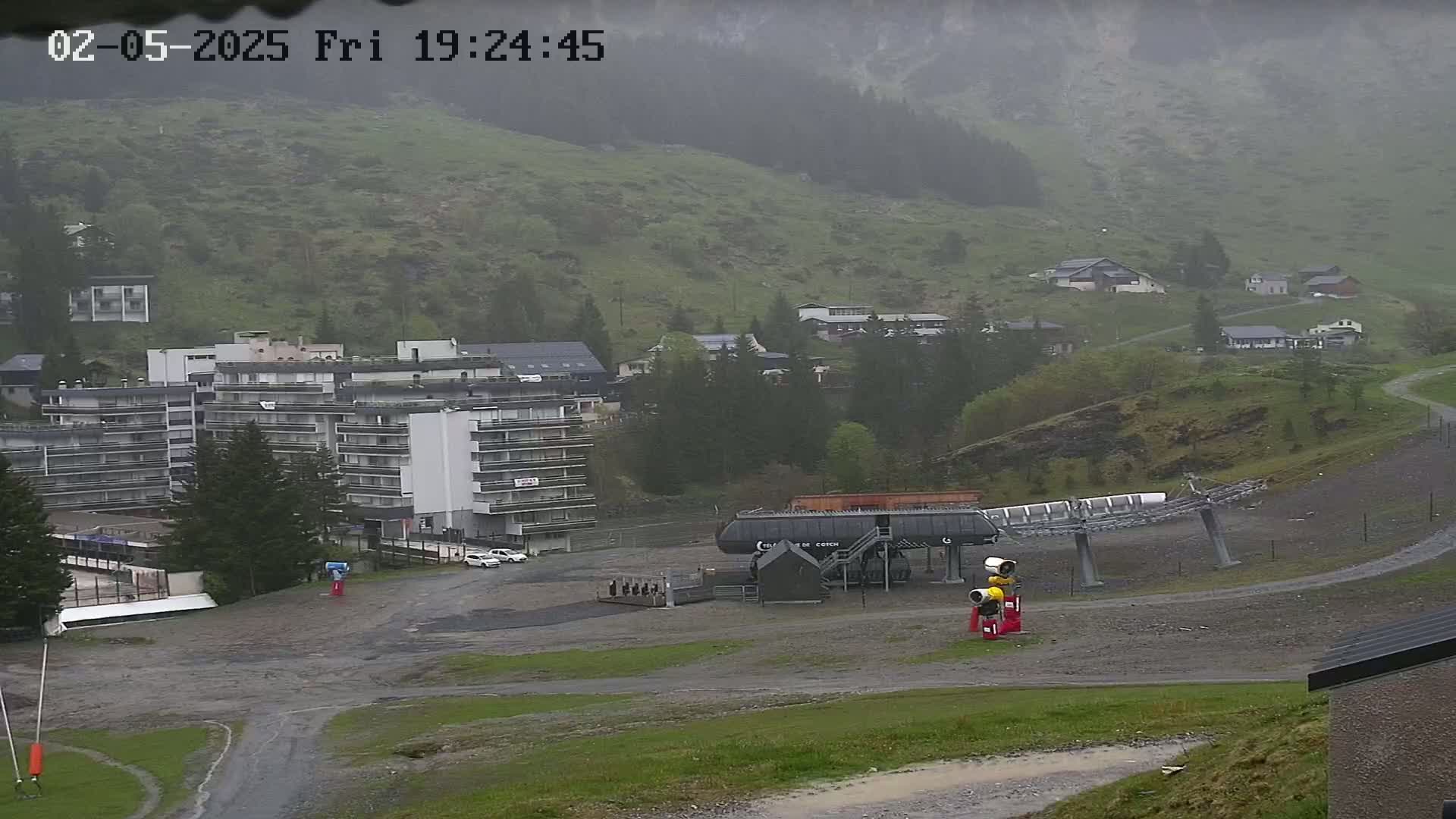 A mountainside village in rainy weather, featuring a large apartment building, a chairlift, and several smaller buildings, all situated on a mostly barren hillside.