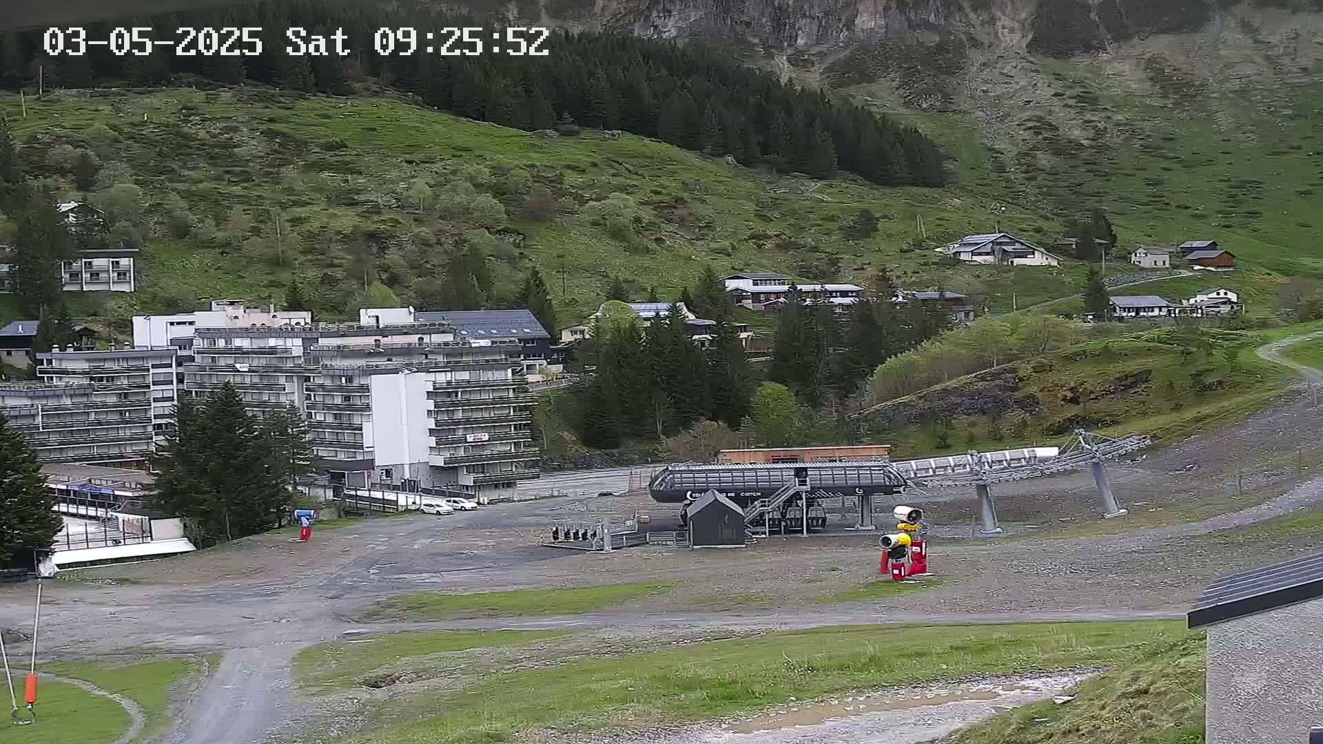 A ski lift sits in a mostly empty gravel lot next to a large apartment building and several smaller buildings on a mountainside under an overcast sky.