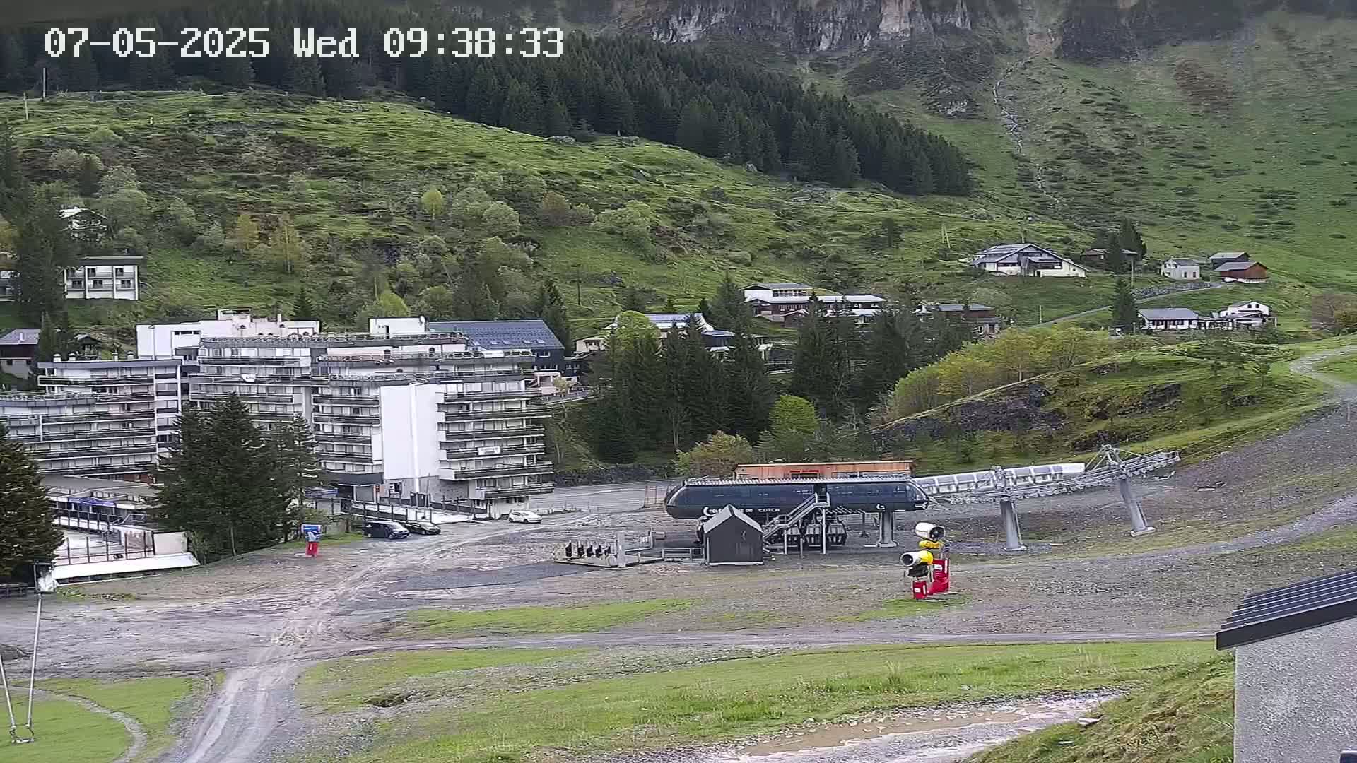 A multi-story building and several smaller houses are situated on a grassy hillside near a chairlift, under overcast skies.