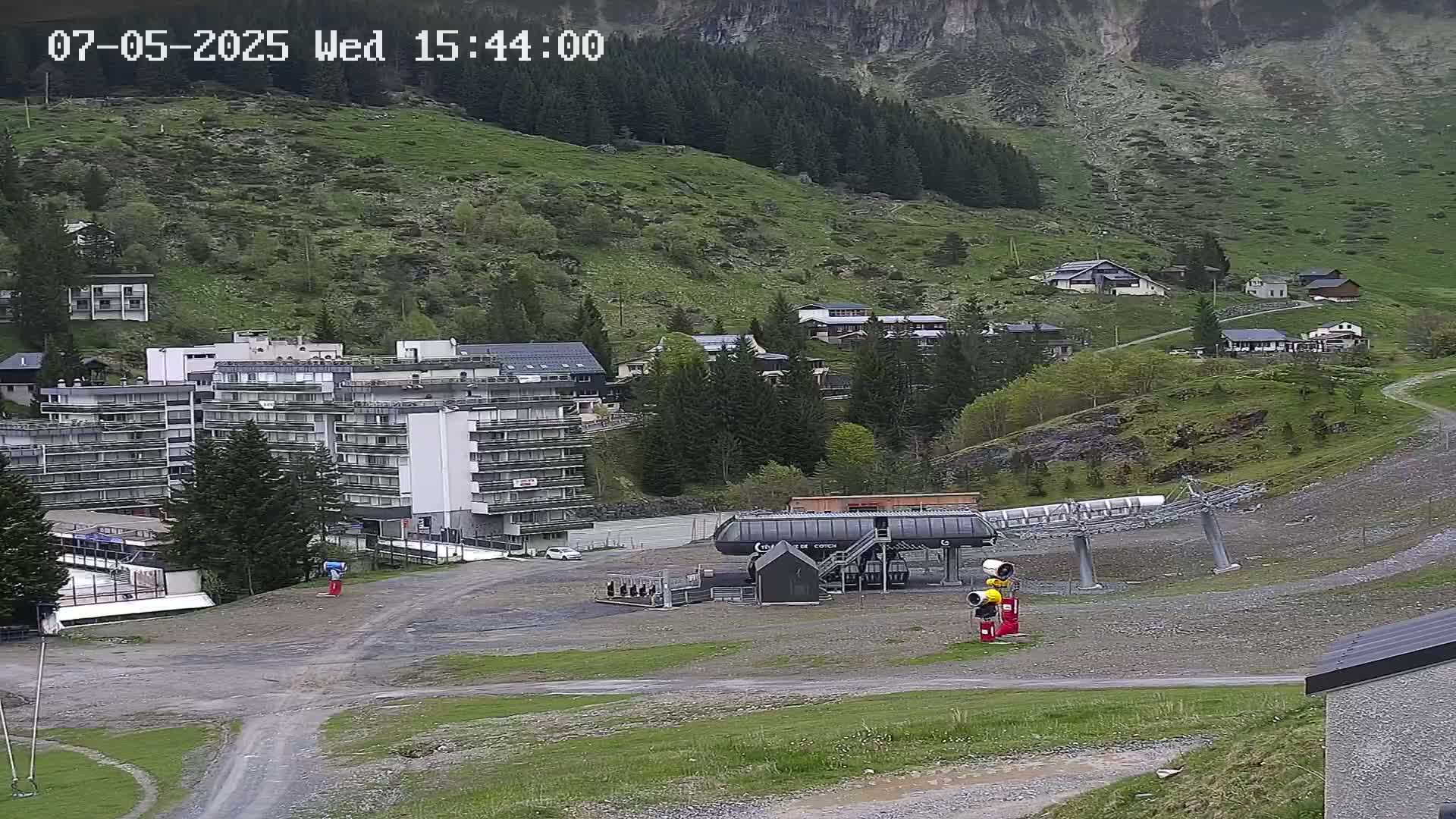 A mountainside village with a large apartment building, a chairlift, and several smaller buildings, under overcast skies.