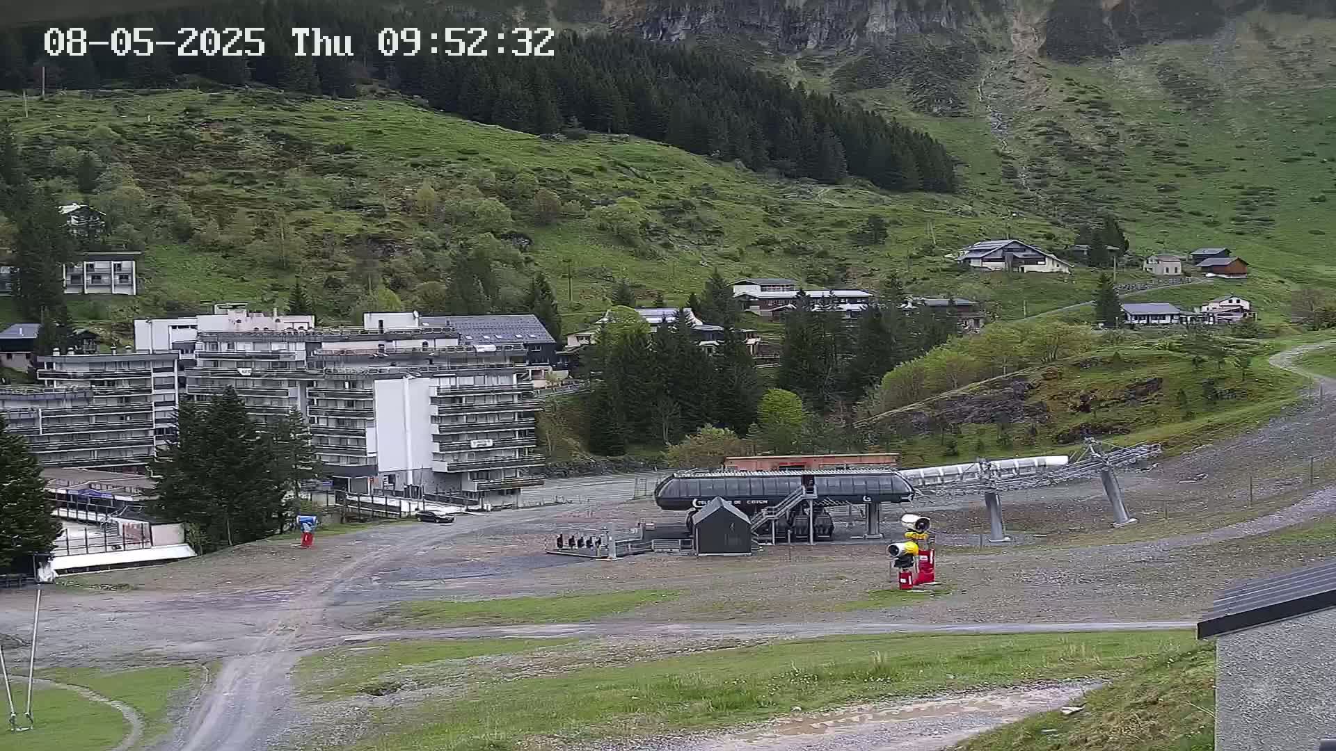 A chairlift base station sits in a gravel area next to a multi-story building in a mountainous village on an overcast day.