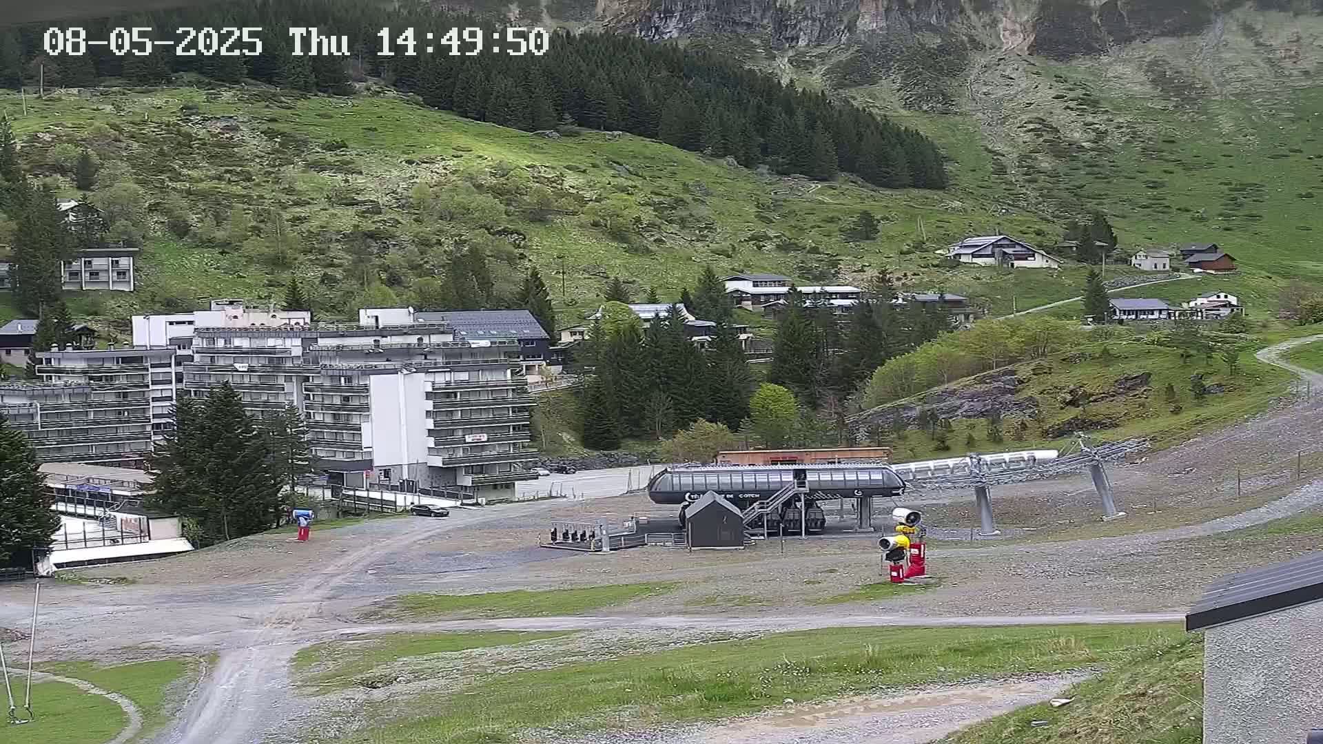 A ski lift base station sits in a mountain village with several buildings, green hillsides, and sparse trees, under overcast skies.