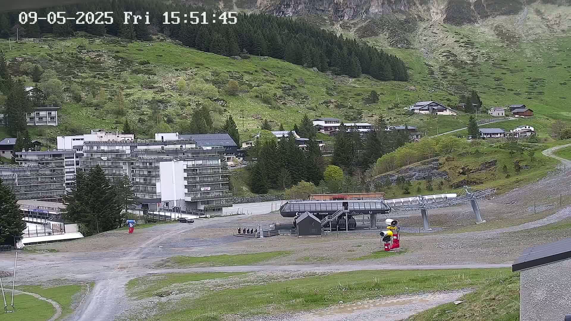 A ski lift base station sits in a gravel lot next to a multi-story building, with other buildings and green hills rising up into the background under an overcast sky.