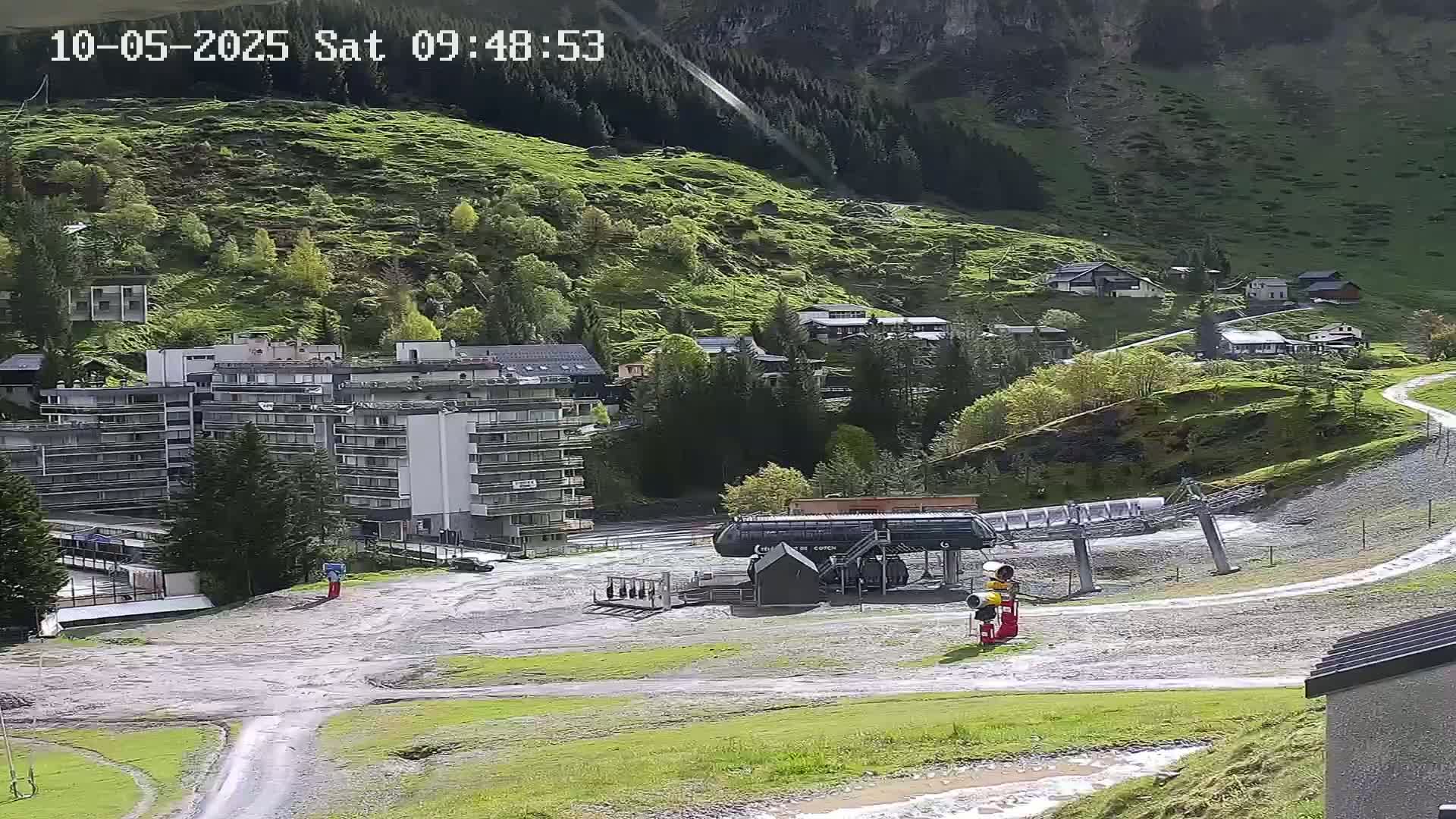 A sunny view of a mountainside village with a multi-story building, a chairlift, and grassy hills.