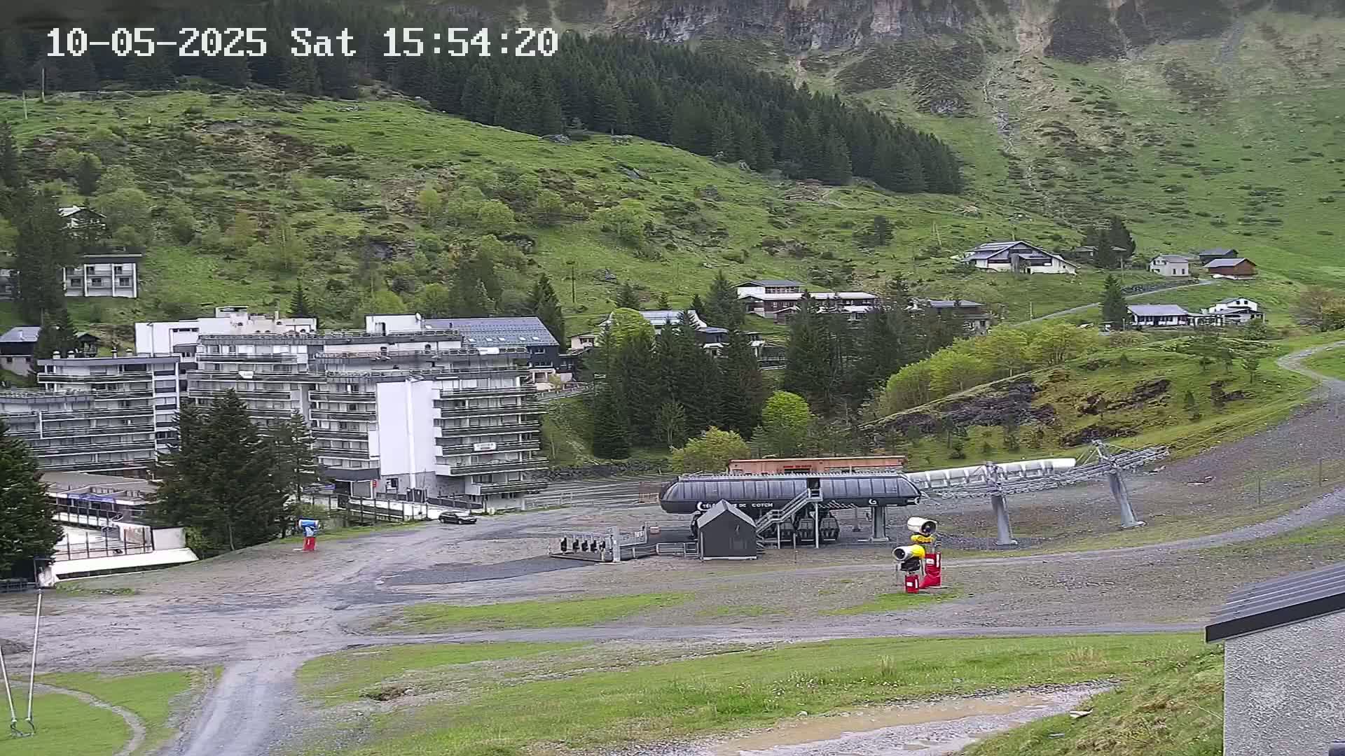 A mountain village with several buildings, including a large apartment complex and a chairlift, is shown on a partly cloudy day.