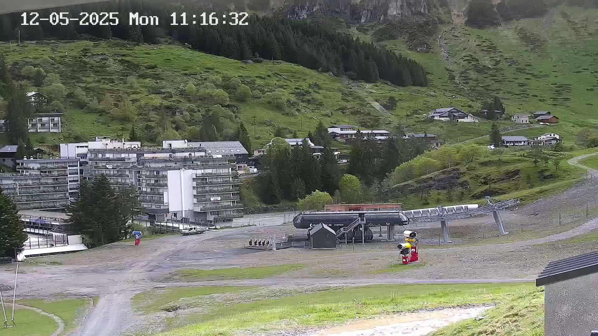 A chairlift base station sits in a mountain village on a mostly overcast day, with a large hotel and several smaller buildings visible on a hillside.