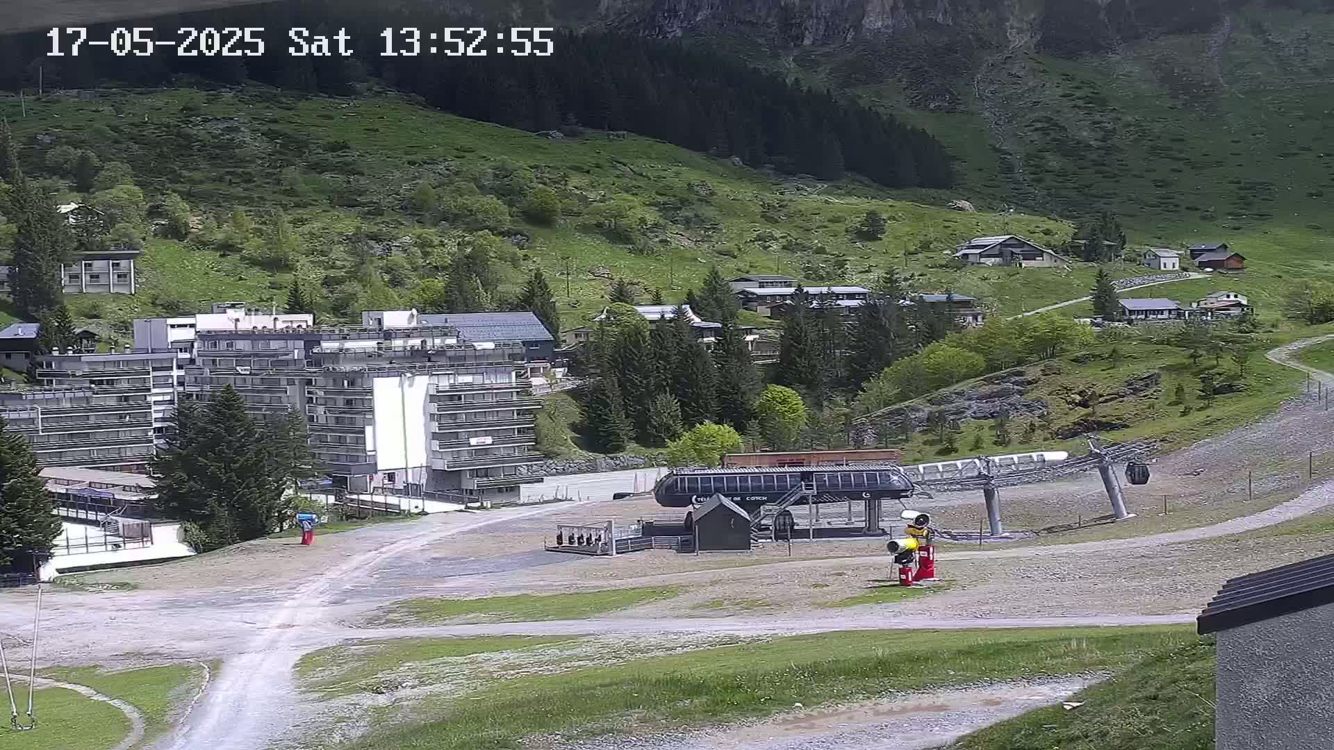 A sunny mountainside village scene shows several buildings, a chairlift, and a grassy hillside under a clear sky.