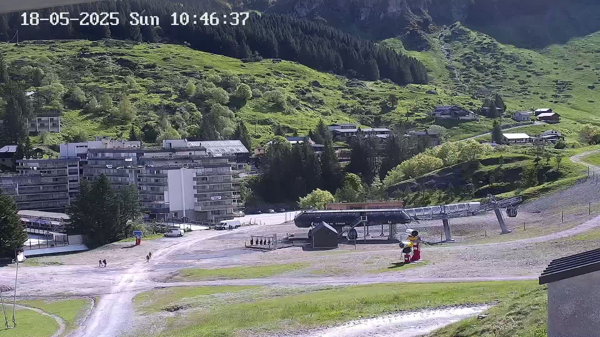 A sunny mountainside village scene shows a chairlift, several buildings, and a few people walking on a dirt path.