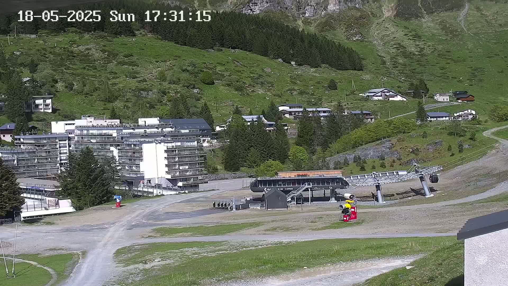 A chairlift base station sits in a grassy area near a multi-story building and several smaller houses nestled into a mountainside on a sunny day.