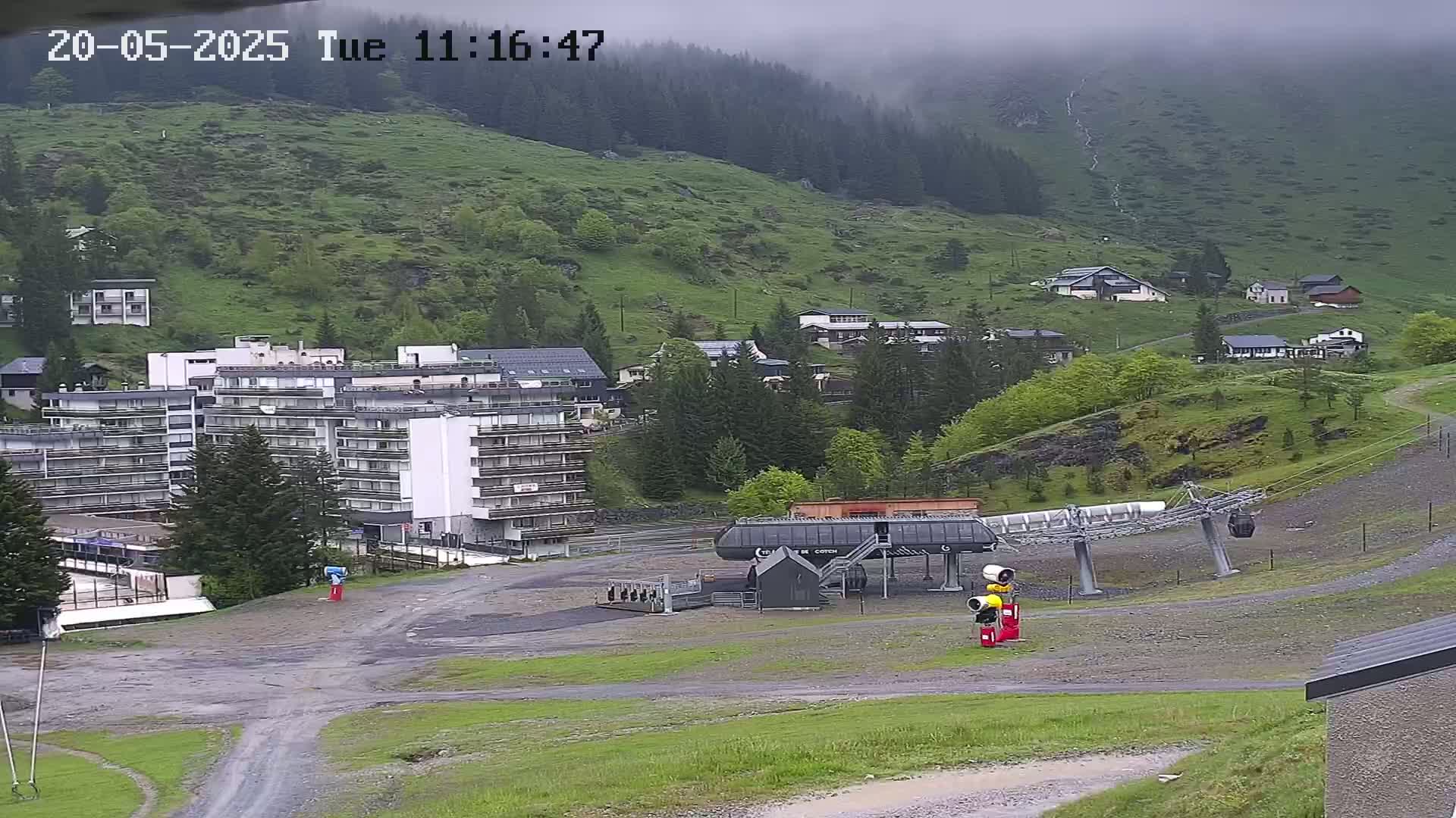 A chairlift base station sits in a valley below a hillside of green grass and trees, under an overcast sky.