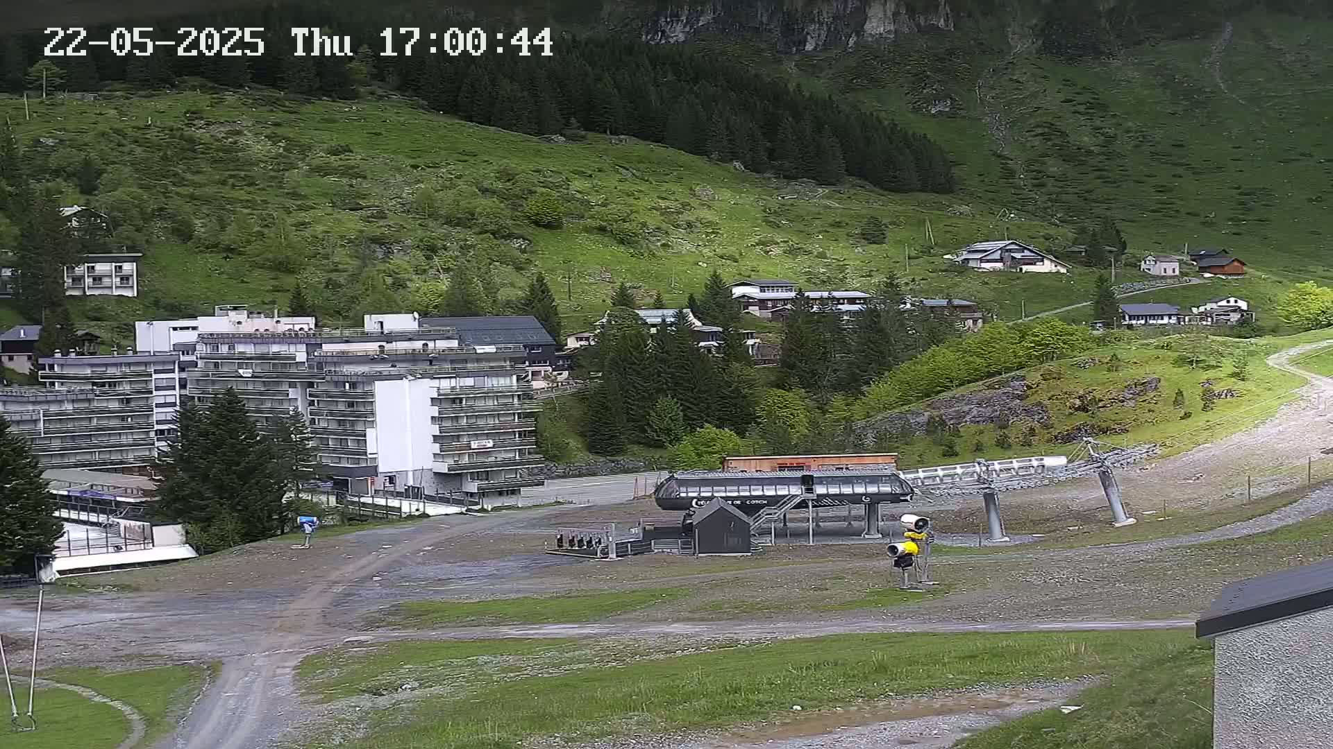 A ski lift base station sits in a mountain valley town on a cloudy day, with several buildings, mostly apartment complexes and smaller houses, visible on the hillside.