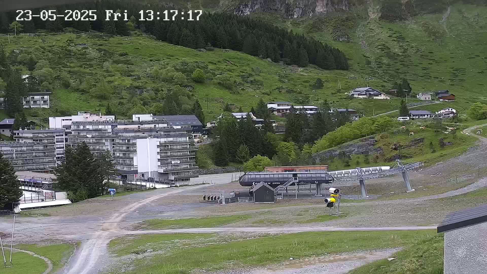 A mountainside village with buildings, a ski lift, and grassy areas under partly cloudy skies.