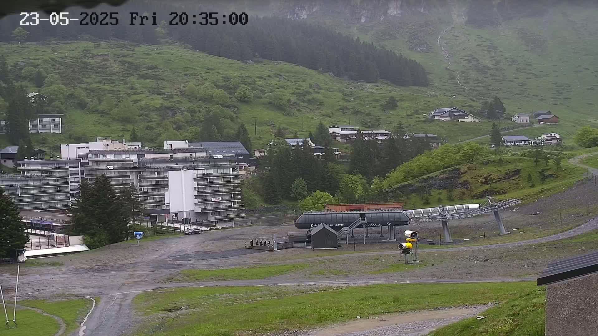 A mountain village is shown on a rainy day, featuring a large apartment building, several smaller homes, and a chairlift base station.