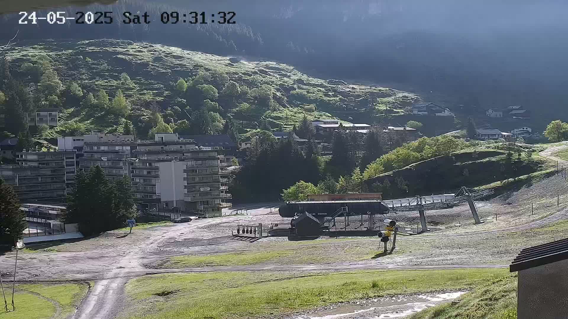 A mostly sunny mountain village scene shows a ski lift, several buildings, and a grassy hillside, with some wet patches of ground indicating recent rain or damp conditions.