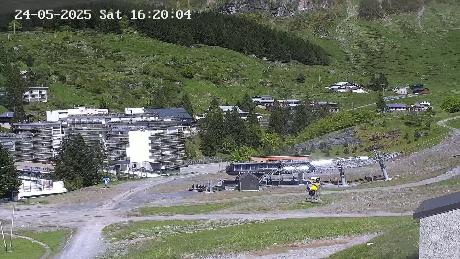 A ski lift base station and several buildings are situated on a grassy hillside under sunny conditions, with a mountain in the background.
