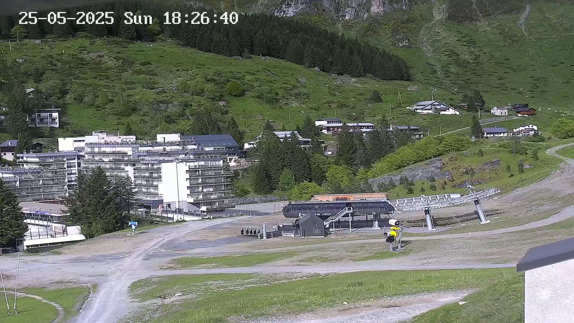 A sunny mountainside village scene shows a large apartment building, several smaller houses, a chairlift, and a mostly empty paved area.